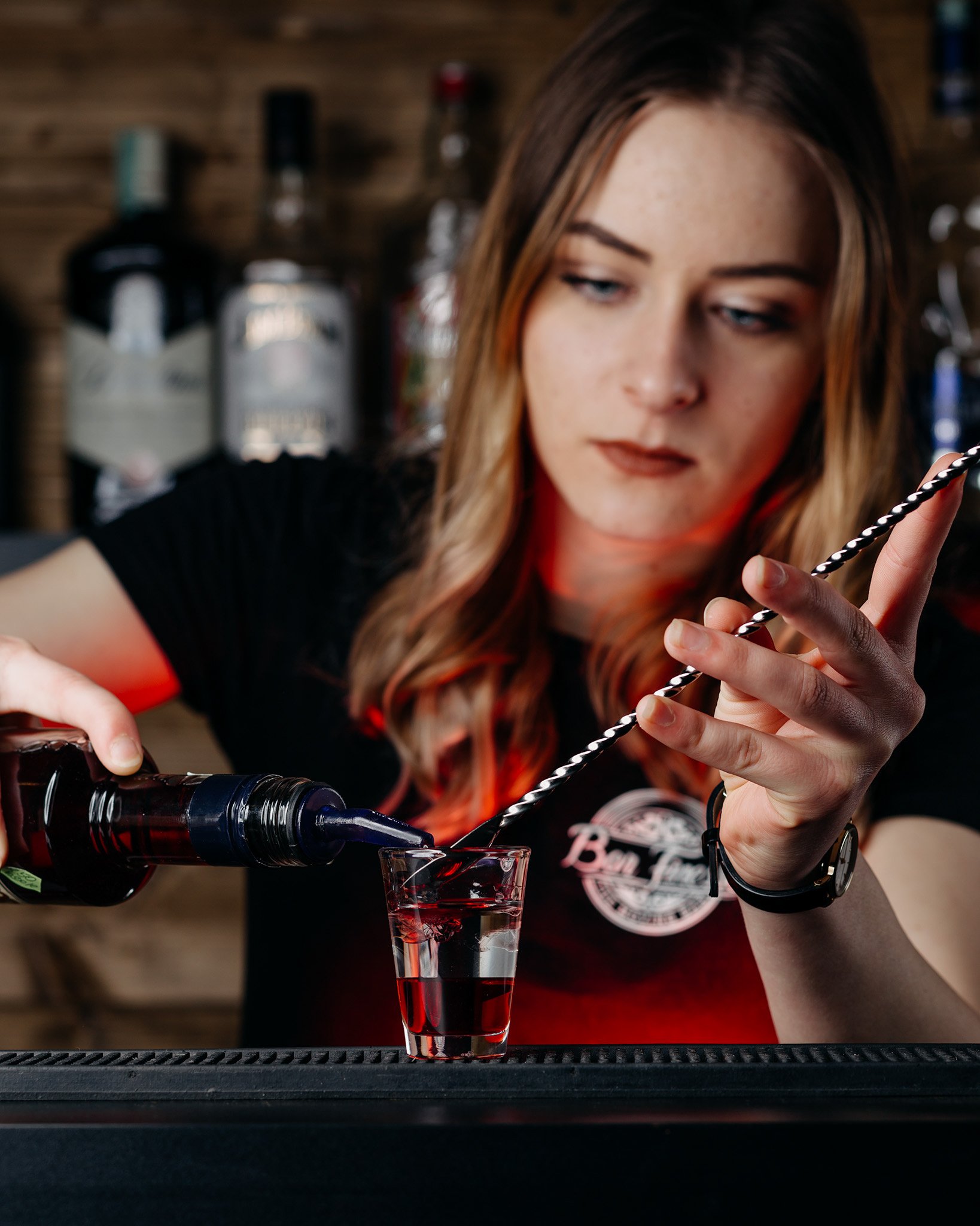 A woman pouring red wine from a bottle into a glass on a bar counter with a wooden wall and liquor bottles in the background.