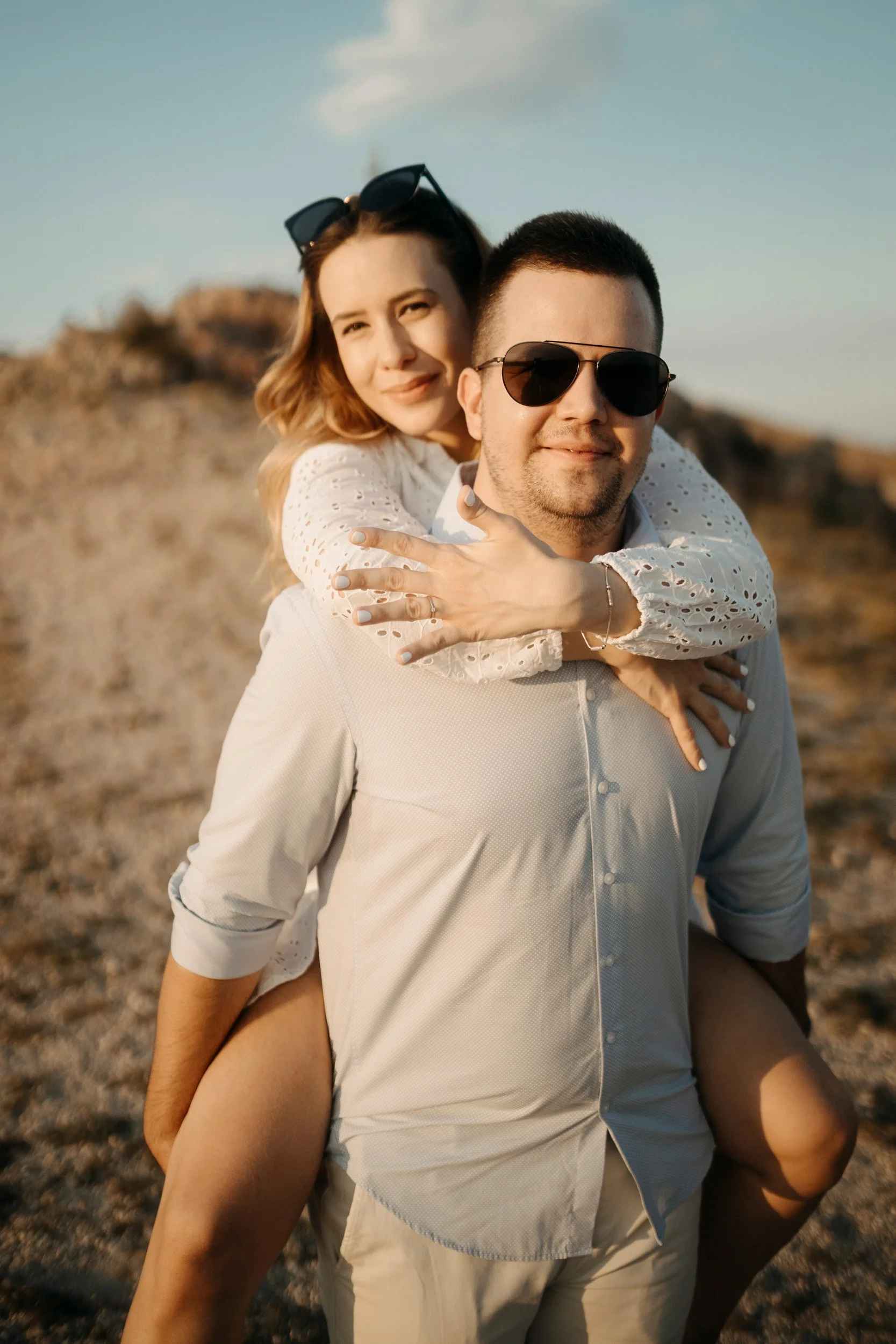 A smiling couple outdoors, with the woman on the man's back, wearing sunglasses. The background is a rocky landscape under a blue sky.