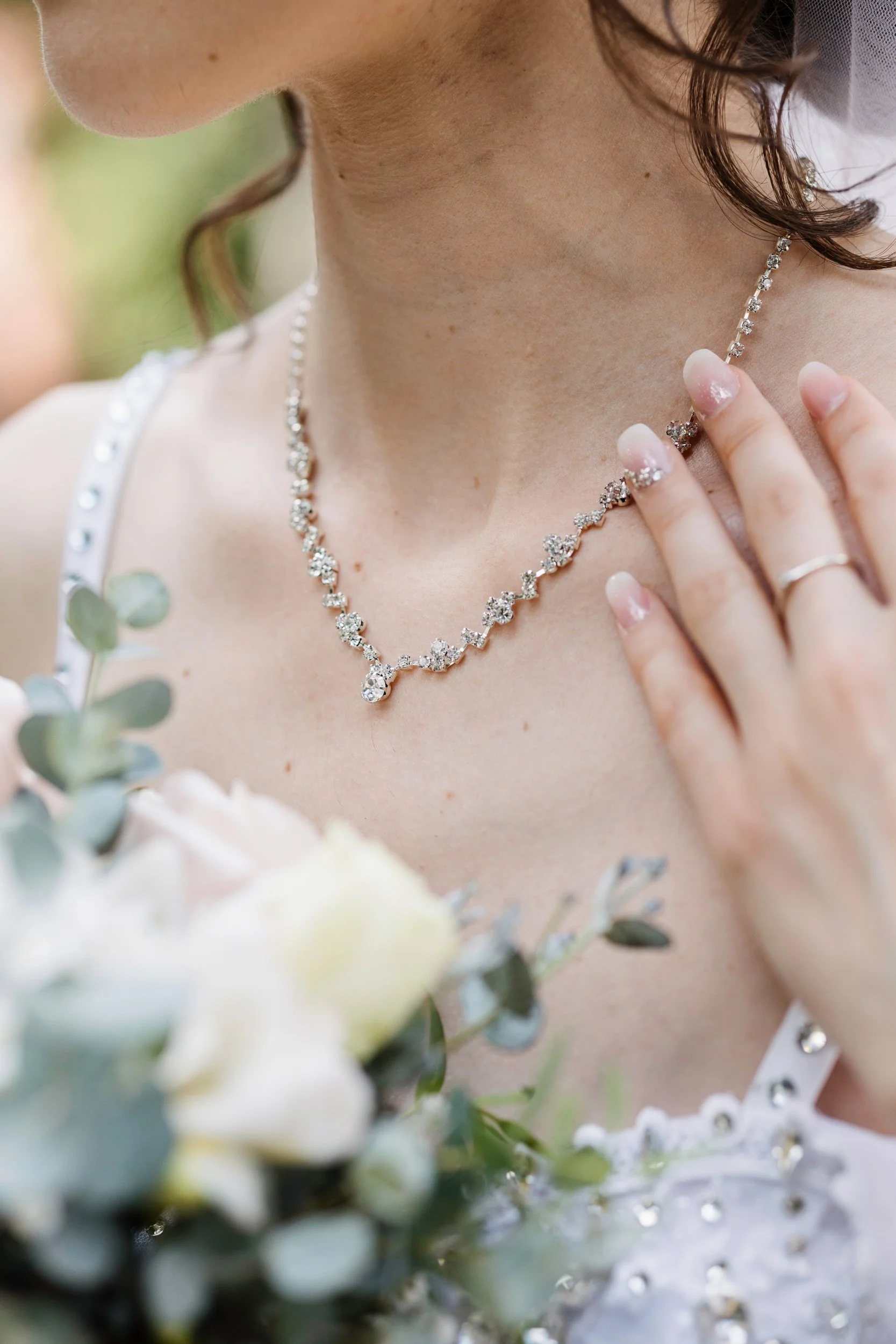 Close-up of a woman wearing a sparkling diamond necklace with her hand touching her neck, dressed in a wedding gown.