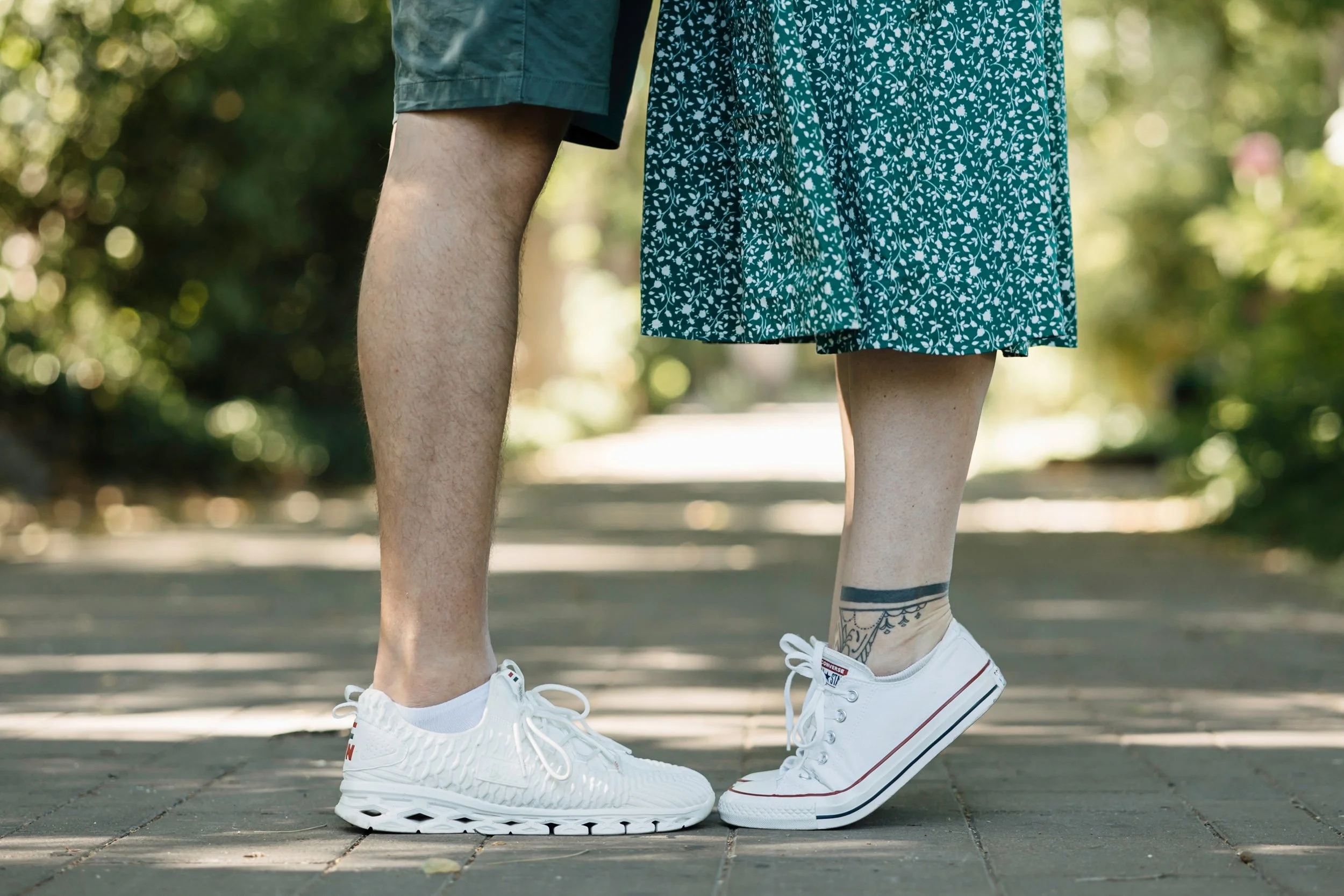 Close-up of a couple's legs and feet standing on a sidewalk, wearing white sneakers with a blurred background of greenery.