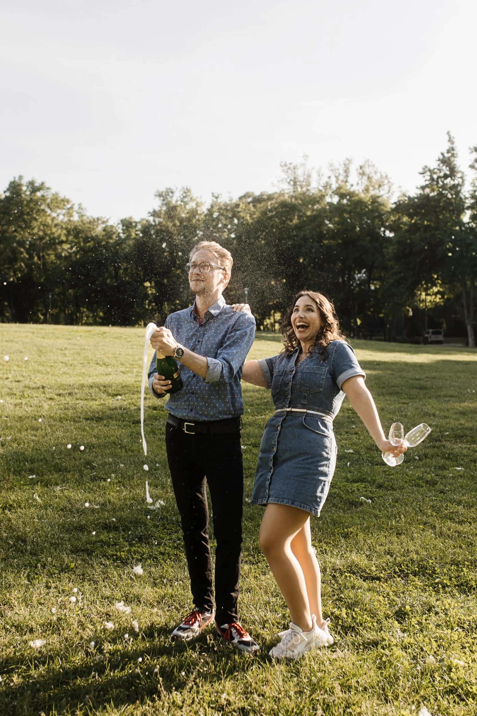 A young man and woman outdoors on a grassy field, playing with champagne and champagne glasses, celebrating, with trees and a clear sky in the background.