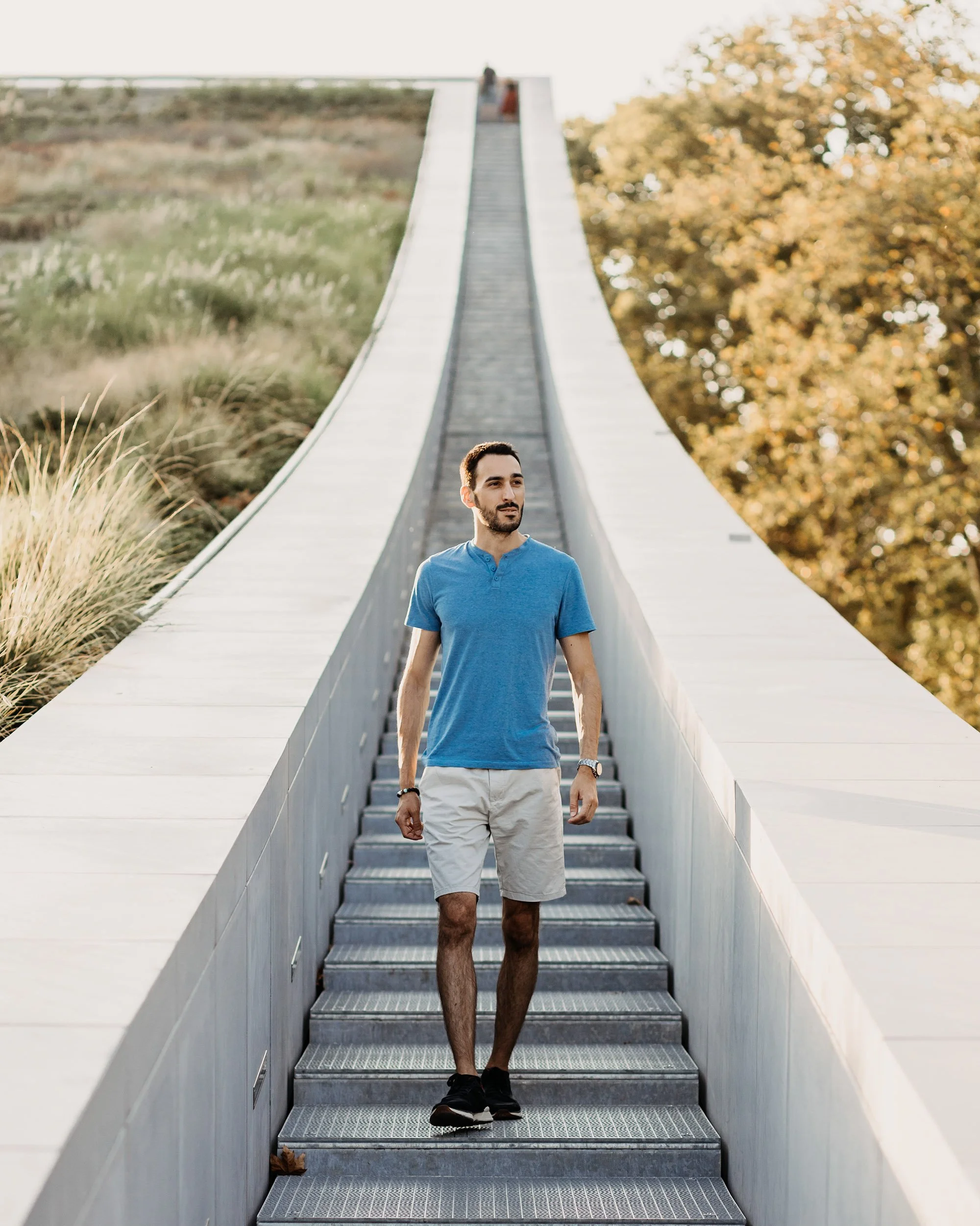 A man in a blue shirt and beige shorts standing on a metal staircase outdoors, with trees in the background.