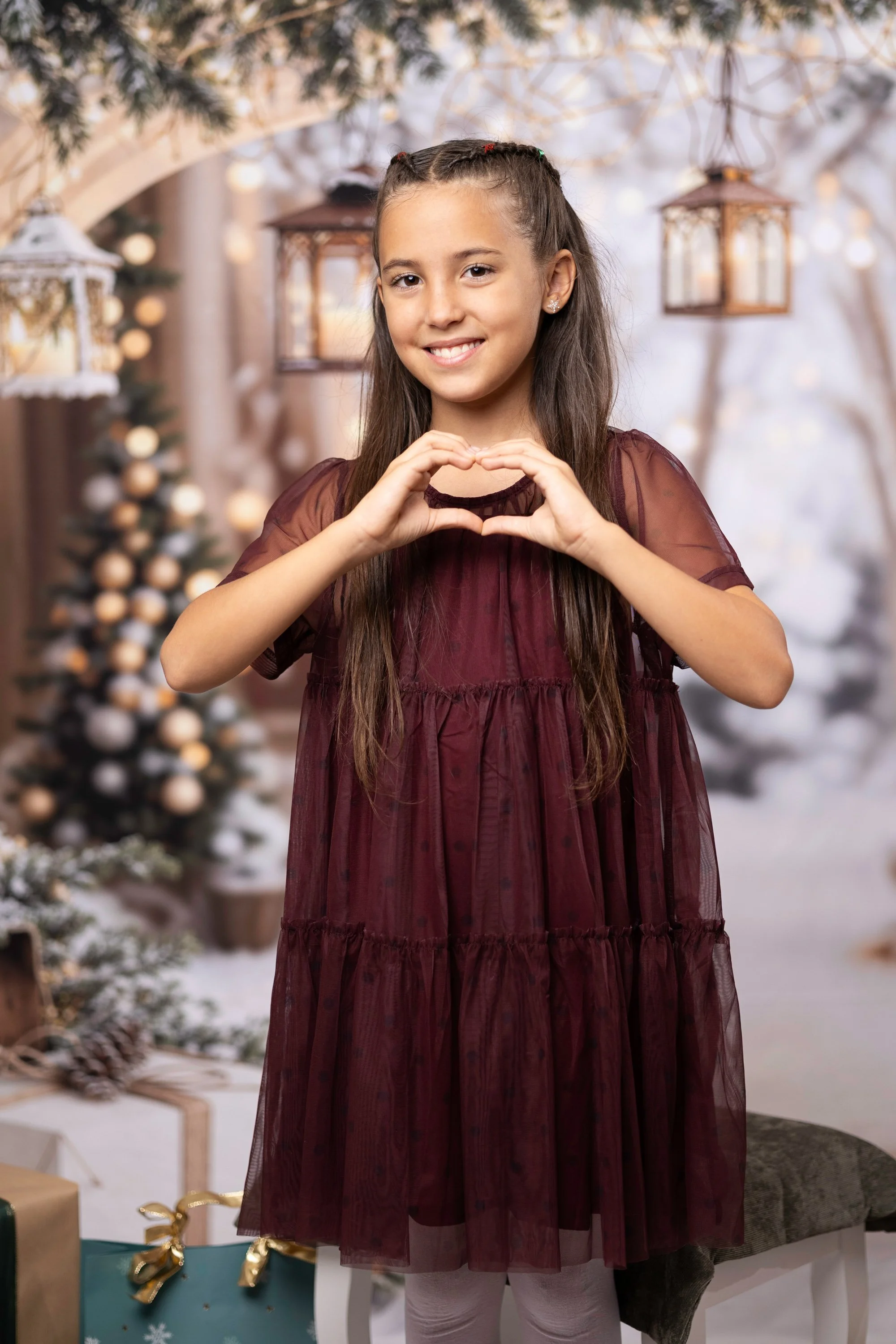 Smiling young girl with long brown hair styled in braids, wearing a burgundy dress and white tights, making a heart shape with her hands in a festive Christmas setting with hanging lanterns, decorated Christmas tree, and wrapped presents.