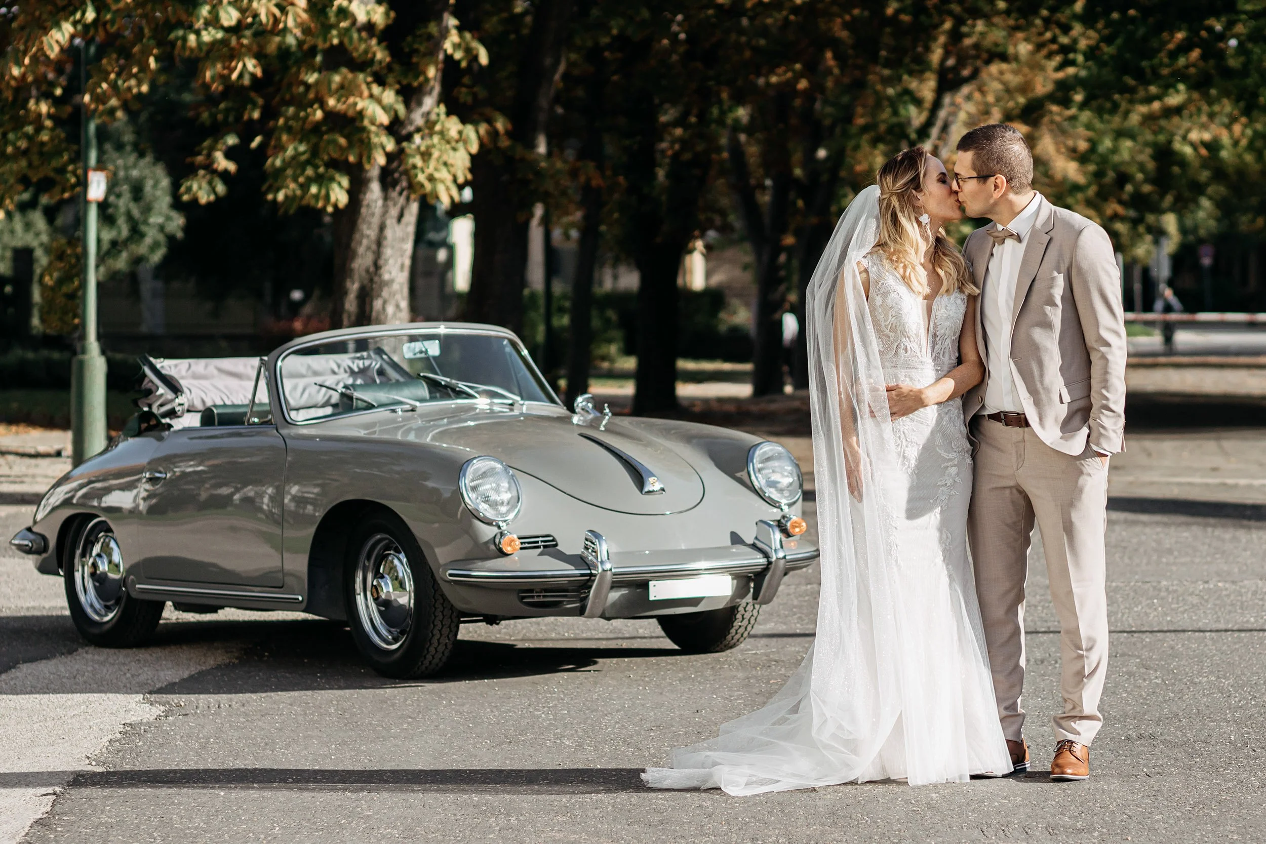A bride and groom kissing next to a vintage silver convertible car in a park with trees in the background.