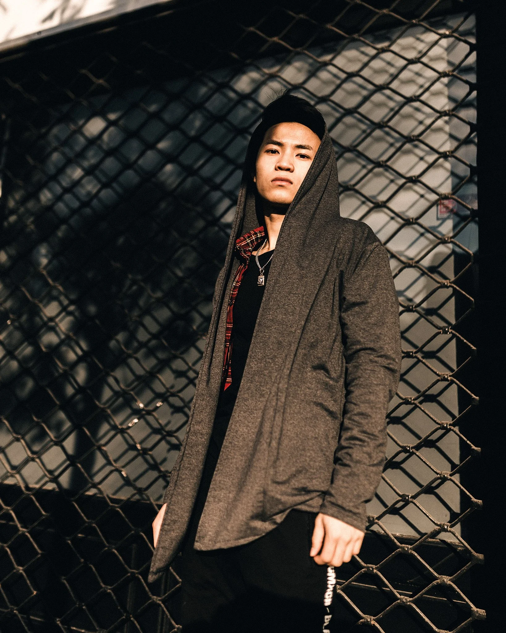 A young man in a hoodie and black pants standing in front of a chain-link fence at night with shadowy buildings in the background.