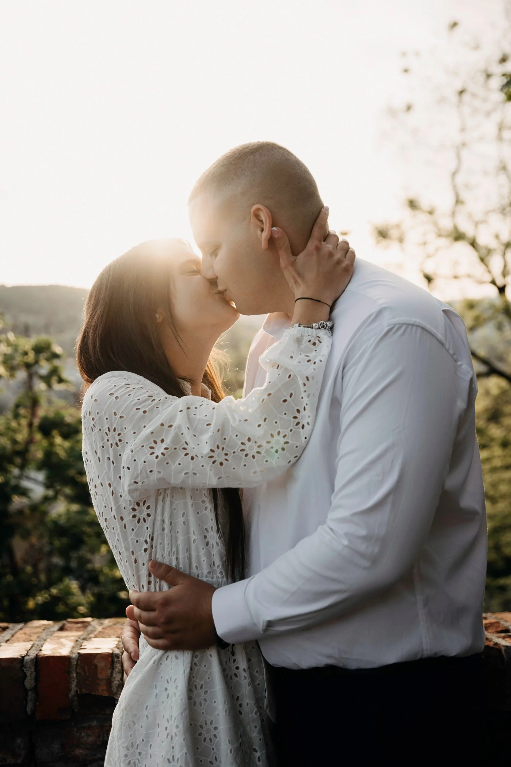 A couple kissing outdoors during sunset, with trees and a brick wall in the background.