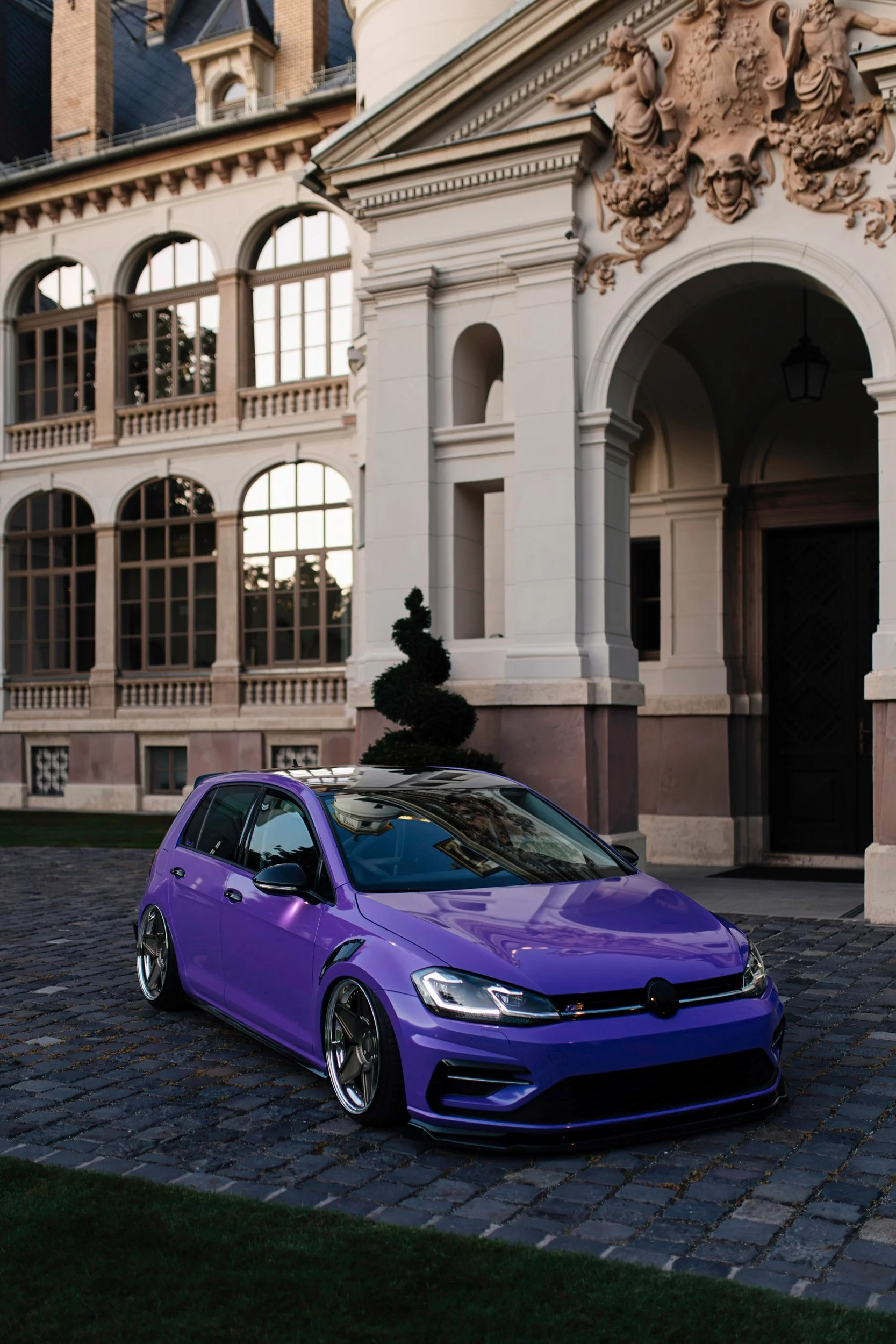A purple sports car parked on a cobblestone street in front of a historic building with ornate architectural details and large windows.