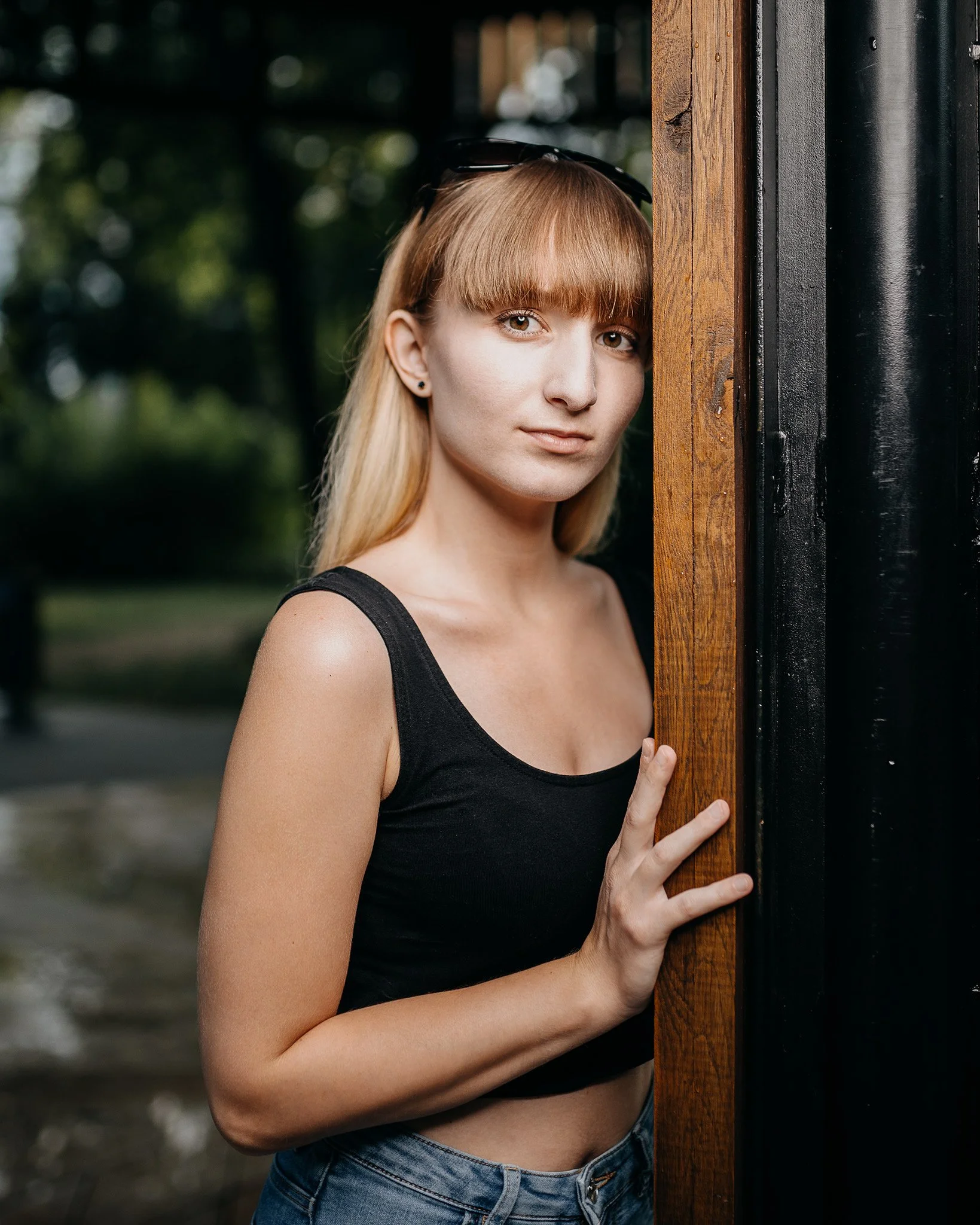 Young woman with straight blonde hair and bangs peeking out from behind a wooden and black panel, outdoors with blurred greenery in the background.