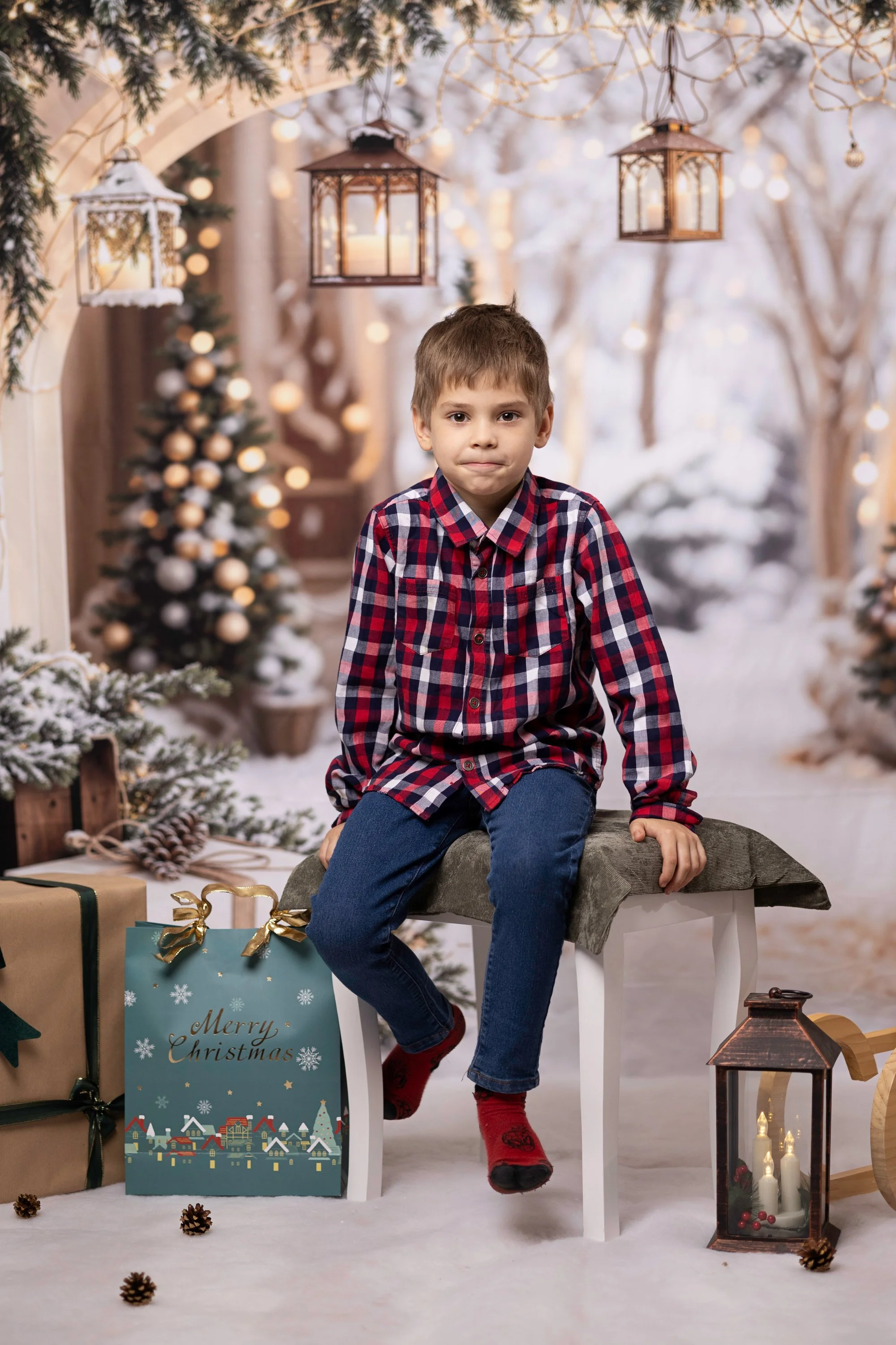 A young boy sitting on a chair in a festive, winter Christmas scene. There are Christmas gifts, pinecones, candles, and a lantern around him. The background shows snow-covered trees and Christmas lights.