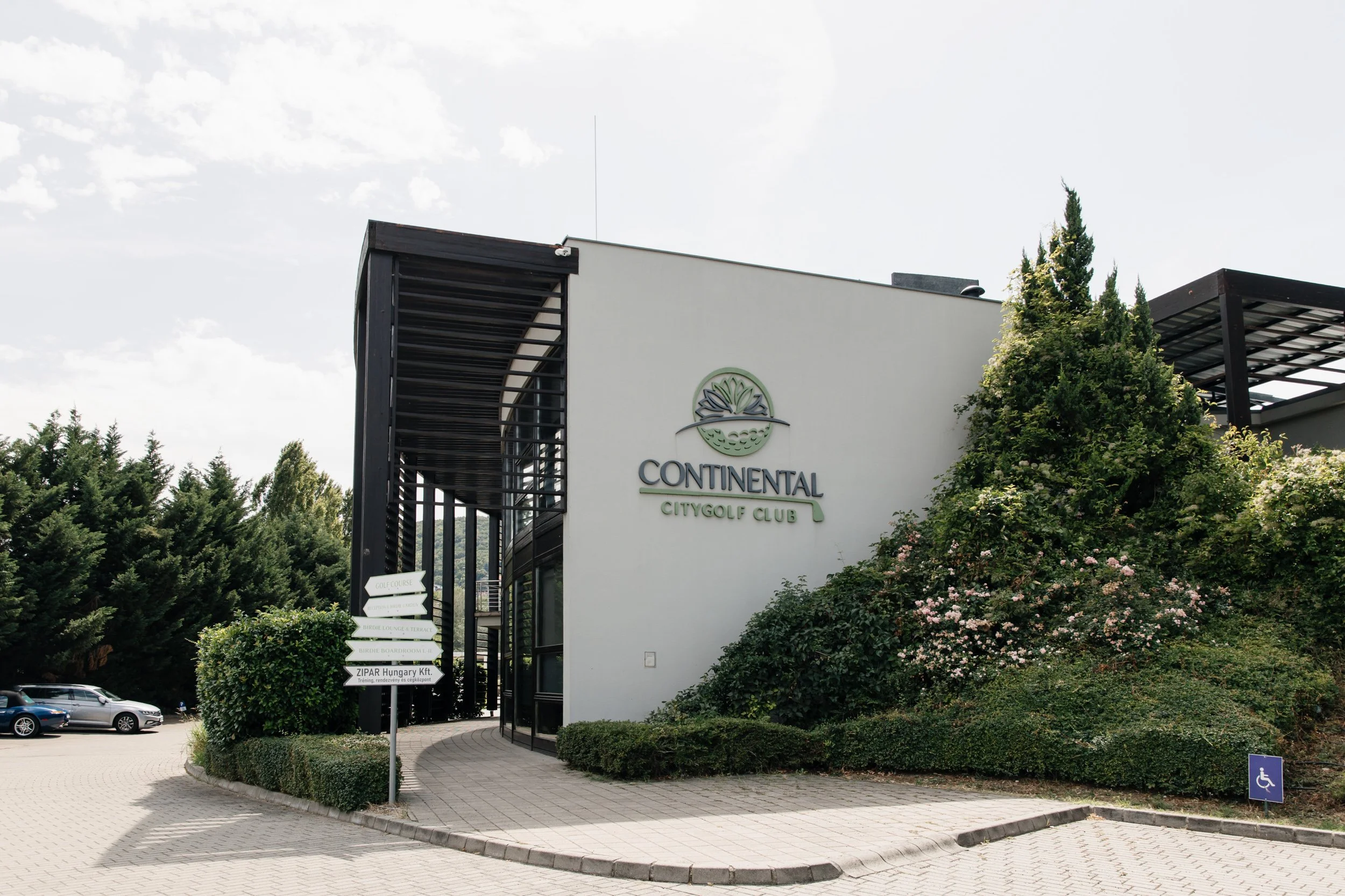 The exterior of the Continental City Golf Club building with a sign displaying various amenities, surrounded by greenery, parking lot, and a clear sky.