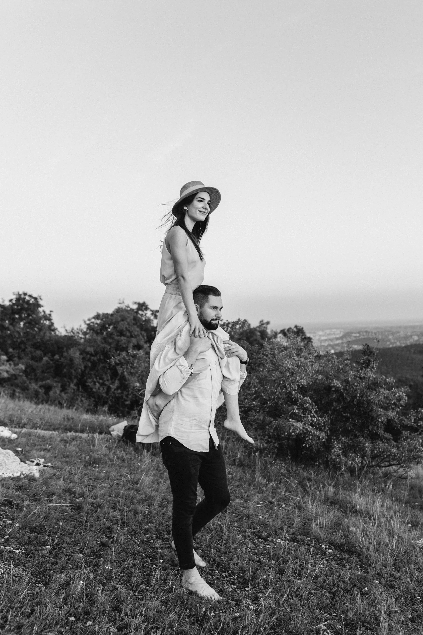A black and white photo of a man carrying a woman on his shoulders. The woman, wearing a hat, is smiling and looking into the distance. They are in an outdoor setting with grassy fields and trees in the background.