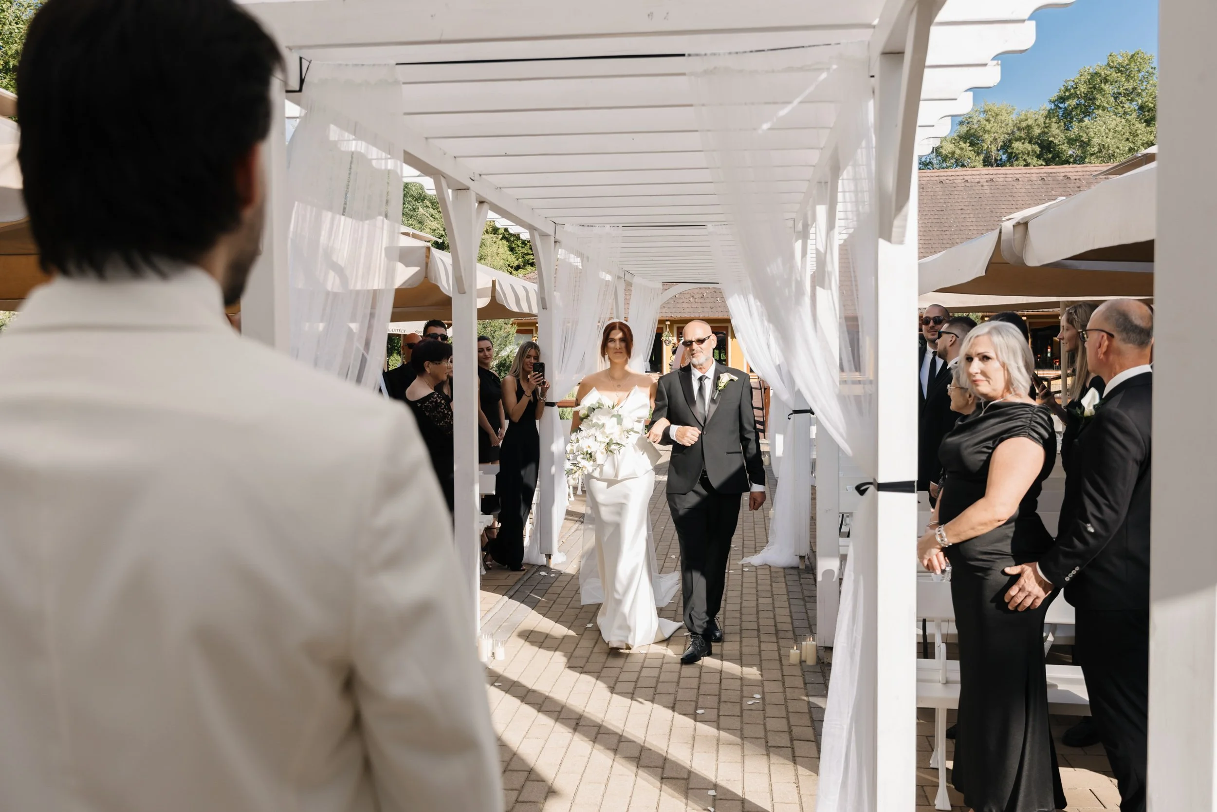Bride walking down the aisle with a man, possibly her father, at an outdoor wedding ceremony surrounded by seated guests.