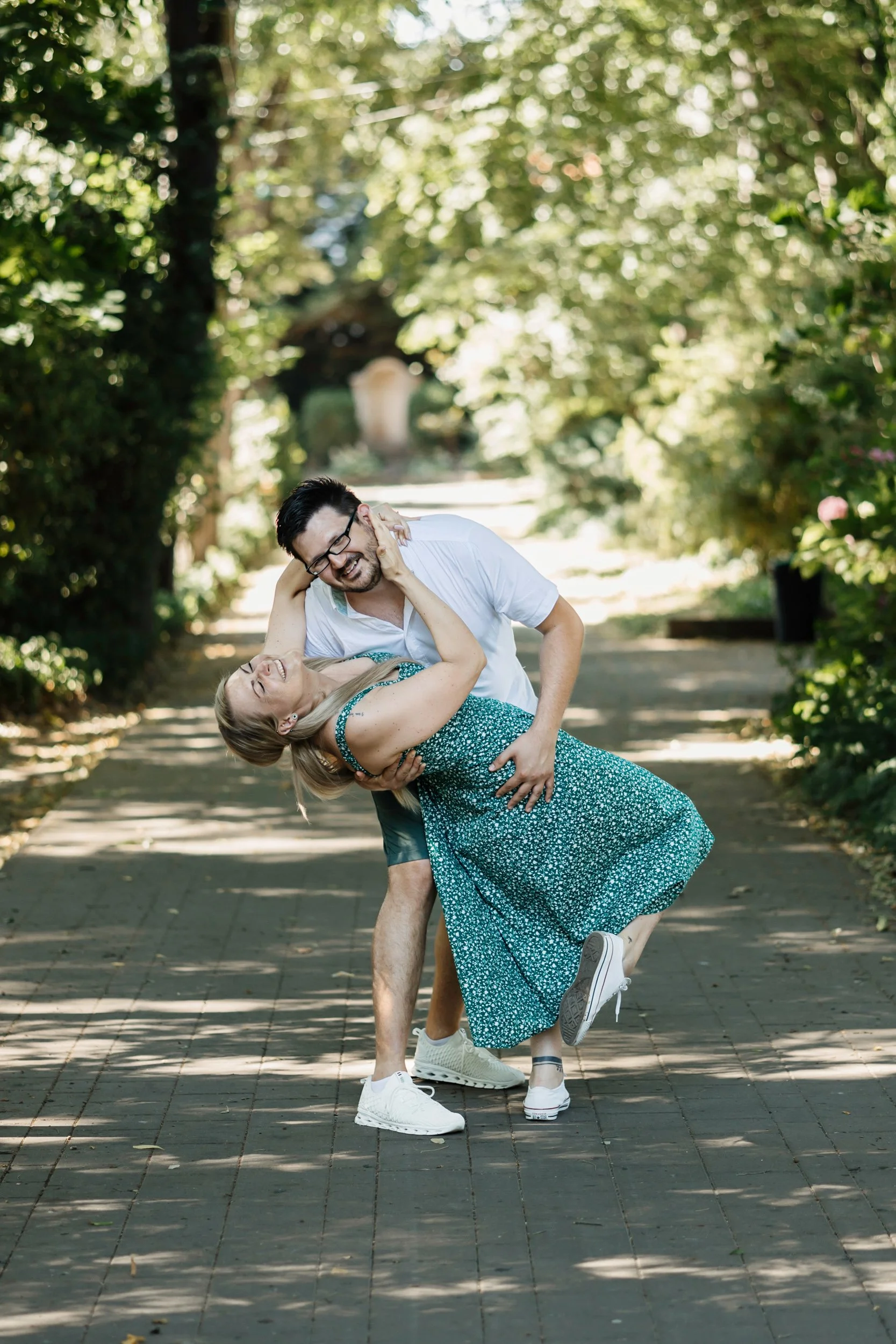 A man with glasses and a beard in a white shirt is holding a woman in a green patterned dress and white sneakers in a dip, both smiling and laughing in a sunny outdoor setting with trees and a walkway.