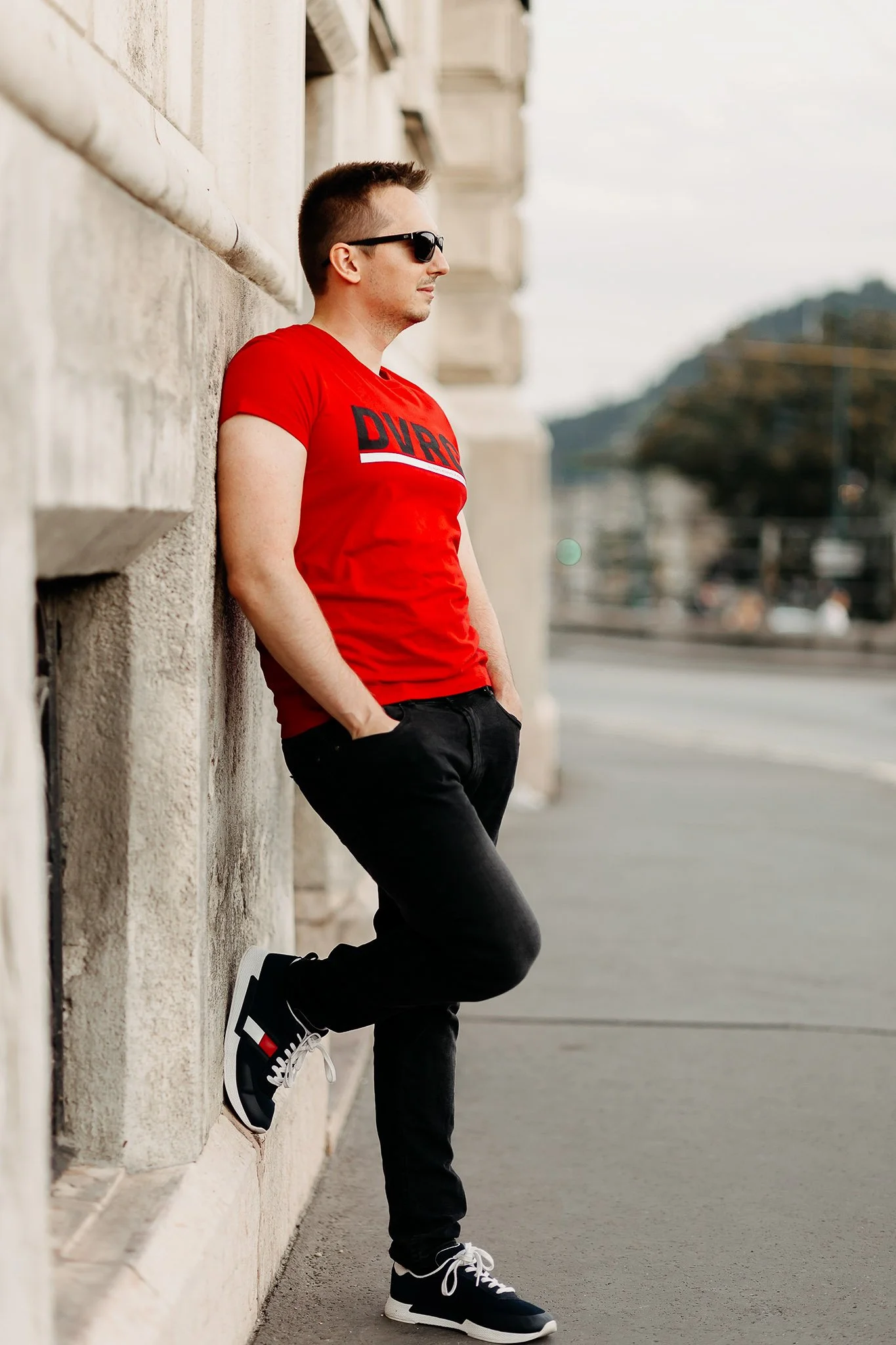 A man wearing sunglasses, a red t-shirt with the word 'DURB', black pants, and black and white sneakers leans against a stone wall with his hands in his pockets and looks to the side.
