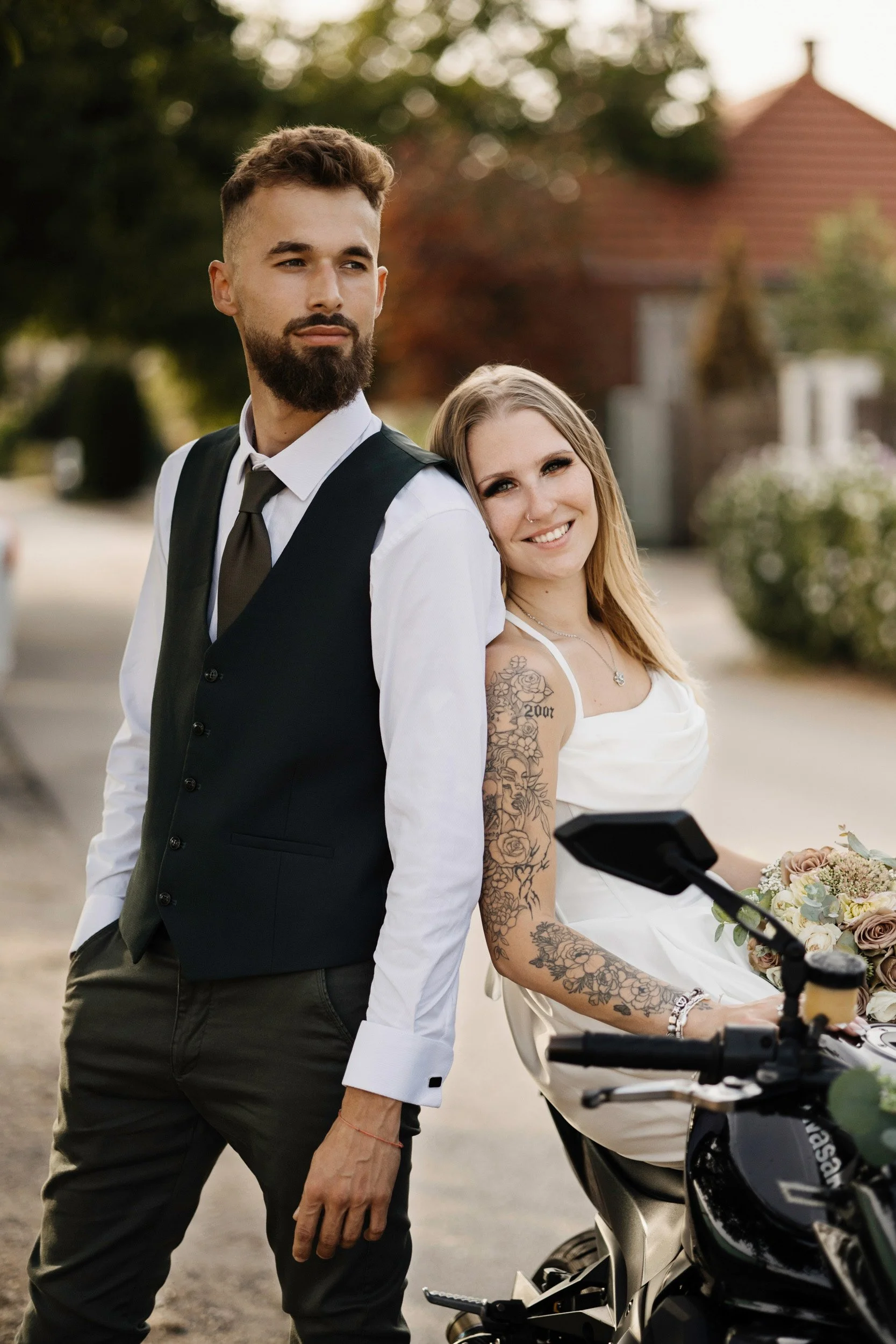 A groom dressed in a white shirt, black vest, and gray pants stands outdoors with a bride who is sitting on a scooter and smiling. The bride, with tattoos and long blonde hair, holds a bouquet of flowers. The background shows trees and a house with a