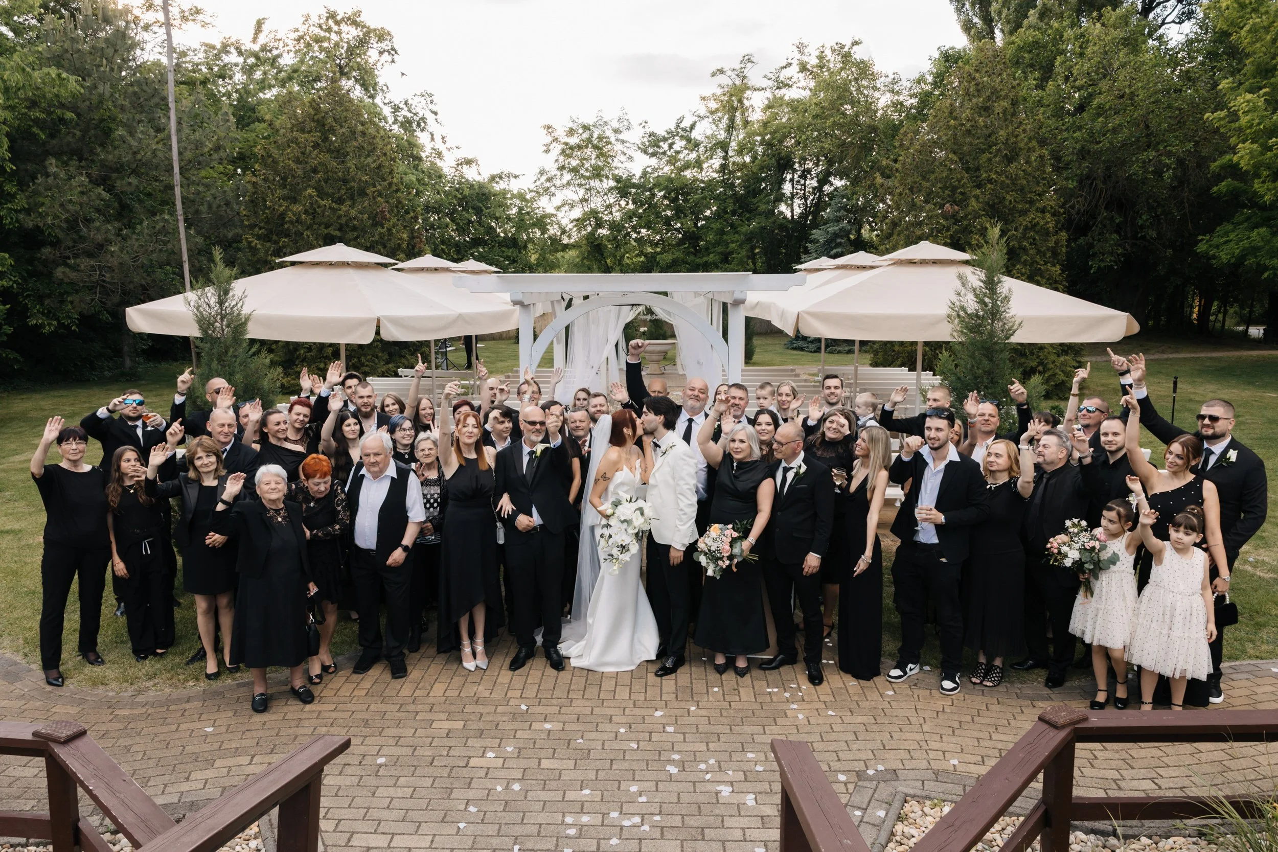 Crowd of wedding guests gathered outdoors, celebrating with a bride and groom in the center, with trees and umbrellas in the background.