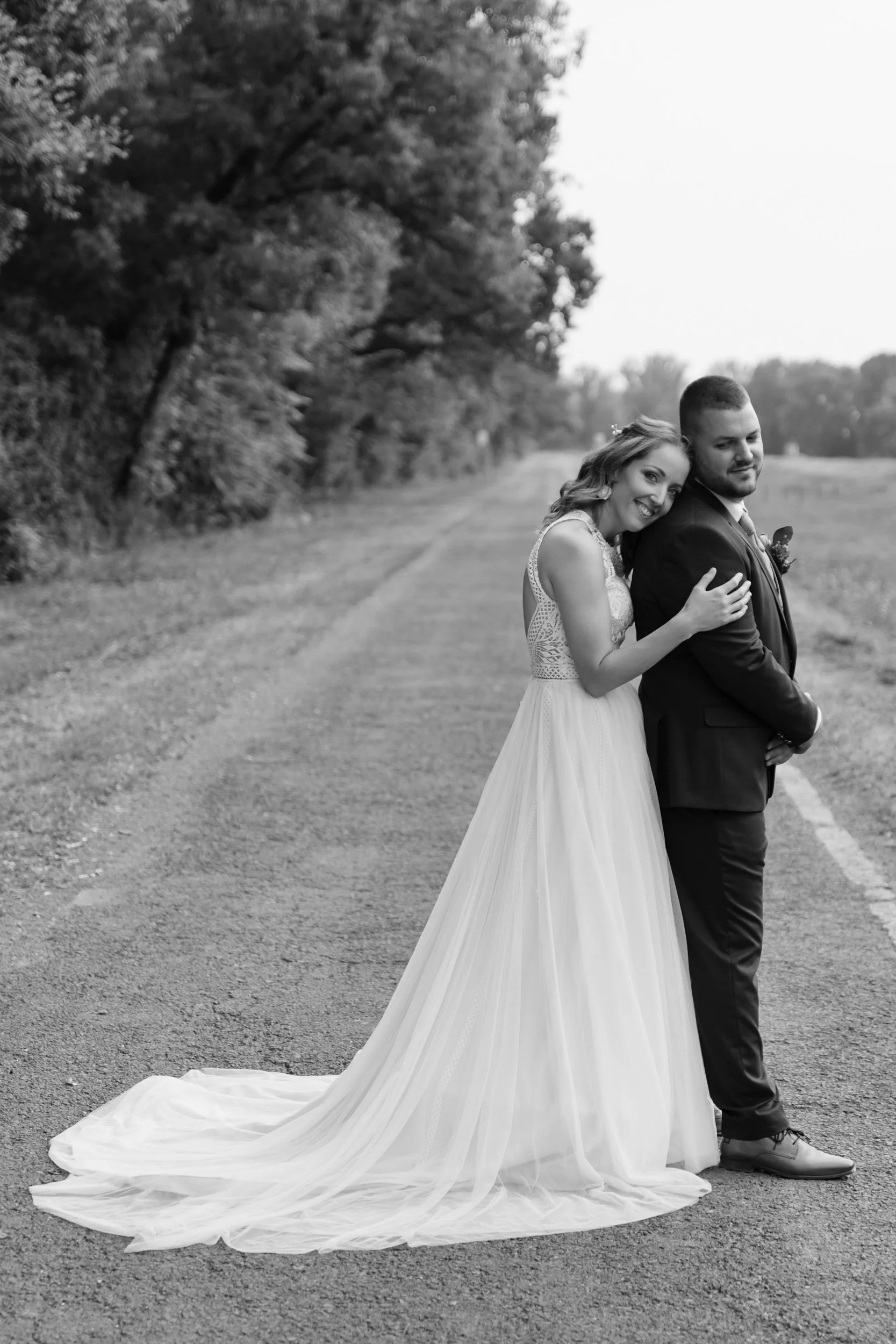 A bride and groom are standing outdoors on a dirt path, embracing each other. The bride is smiling, wearing a lace wedding dress with a flowing train, and leaning her head on the groom's shoulder. The groom is dressed in a suit, with his hands crosse