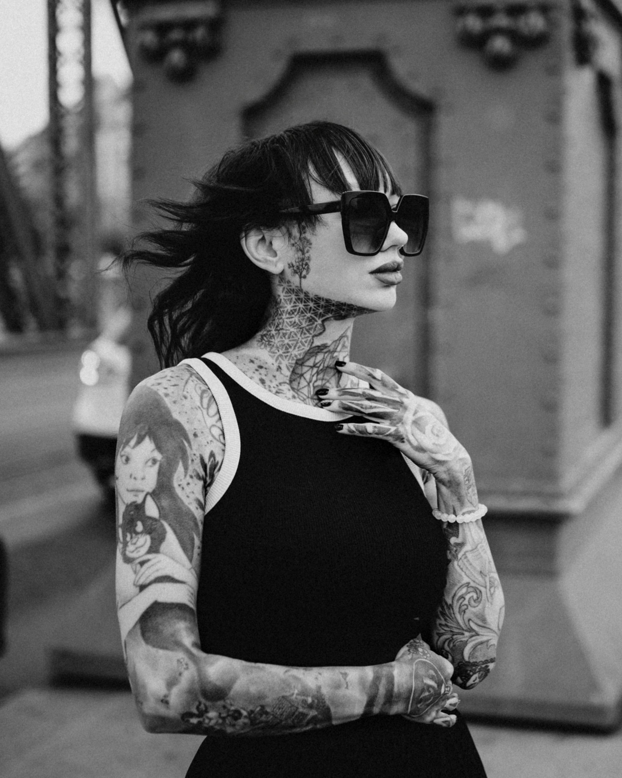 Black and white photo of a woman with tattoos on her arms and neck, wearing large sunglasses, a sleeveless top, and a beaded bracelet, standing outdoors with a blurred background.