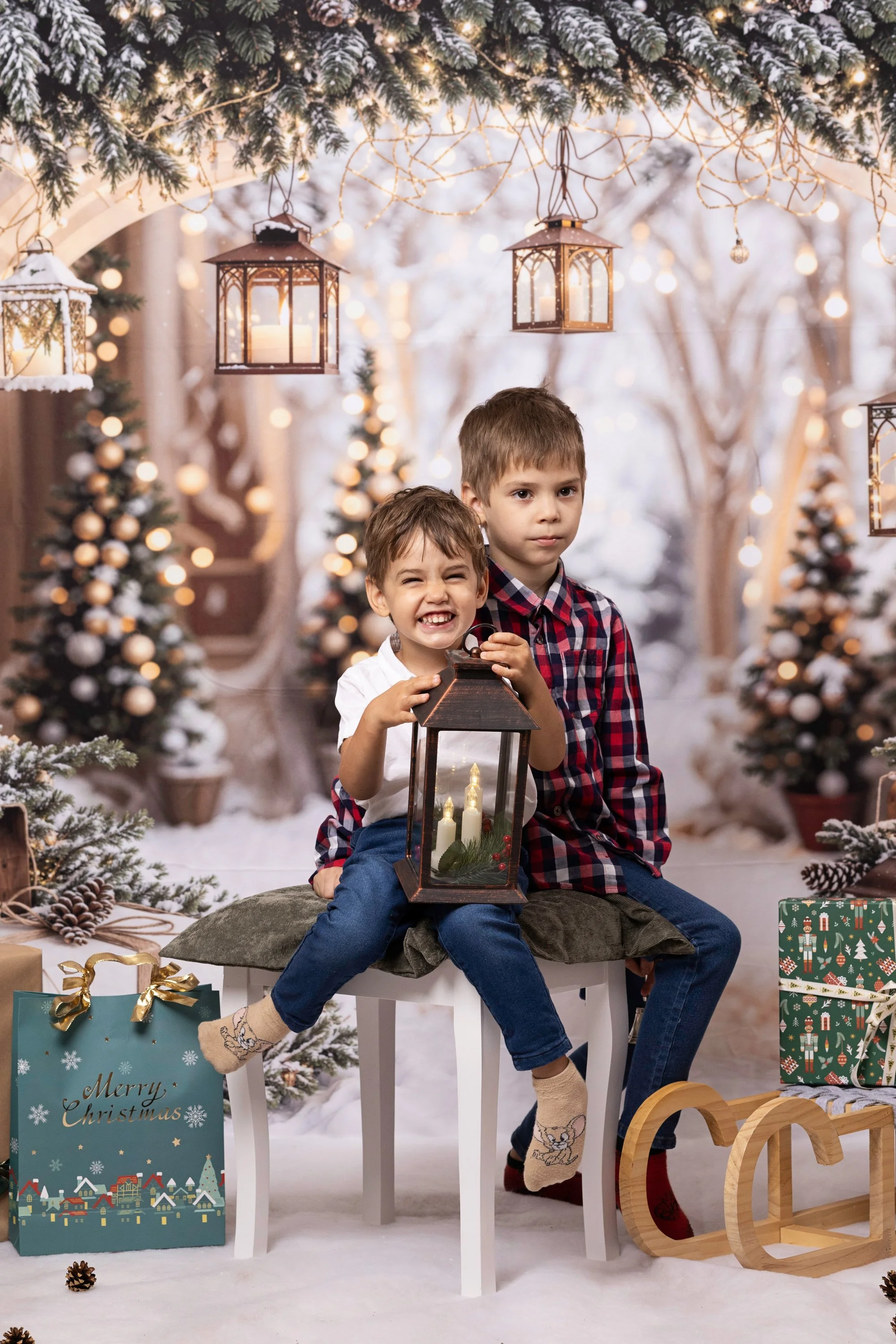 Two young boys sitting on a stool in front of a festive, snowy Christmas scene with decorated trees, hanging lanterns, and holiday presents. One boy smiling, holding a lantern, and the other standing, looking serious.