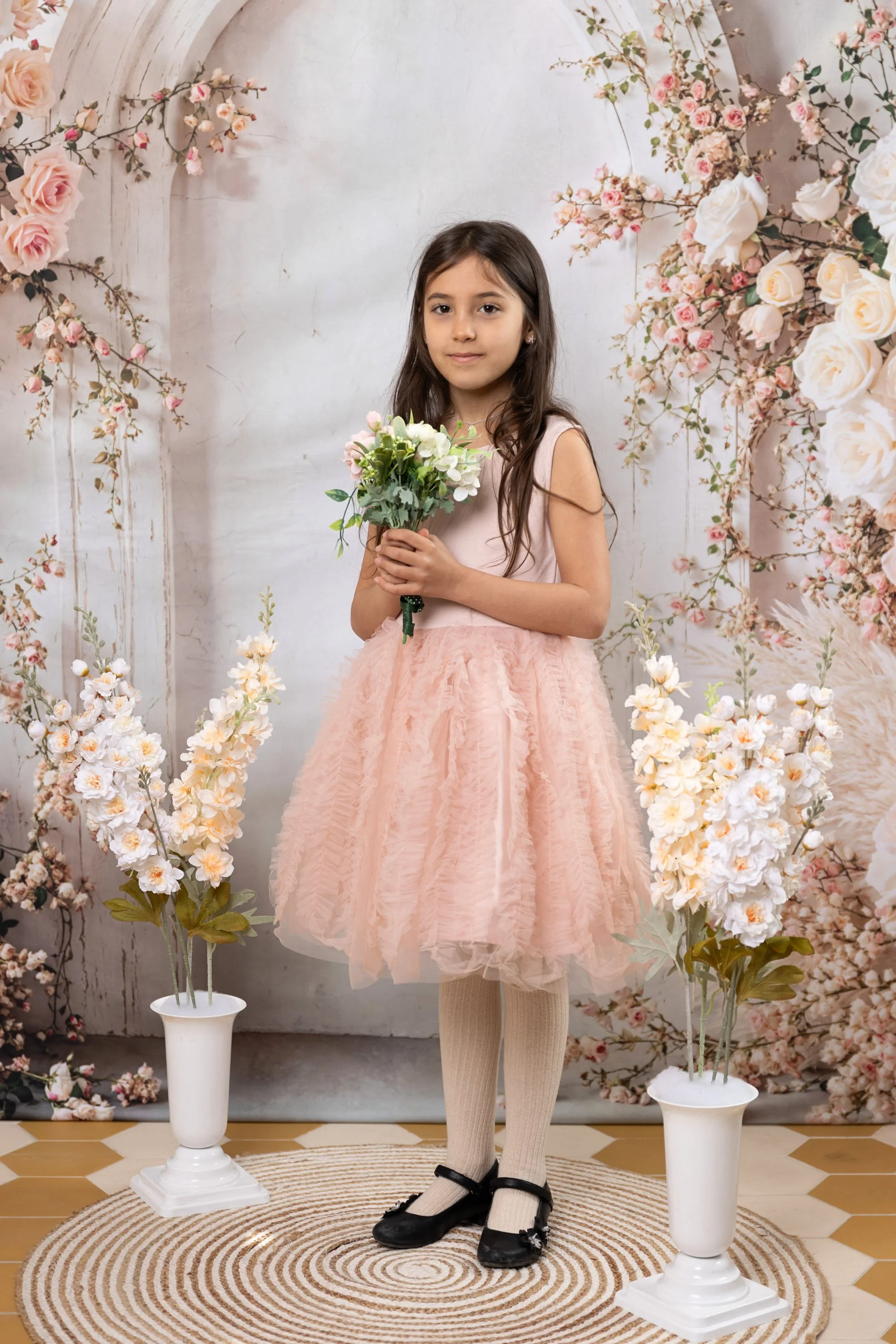 Young girl in pink dress holding a bouquet of white and pink flowers, standing on a circular rug between two white vases with white and pink artificial flowers, decorated with pink and white floral backdrop.