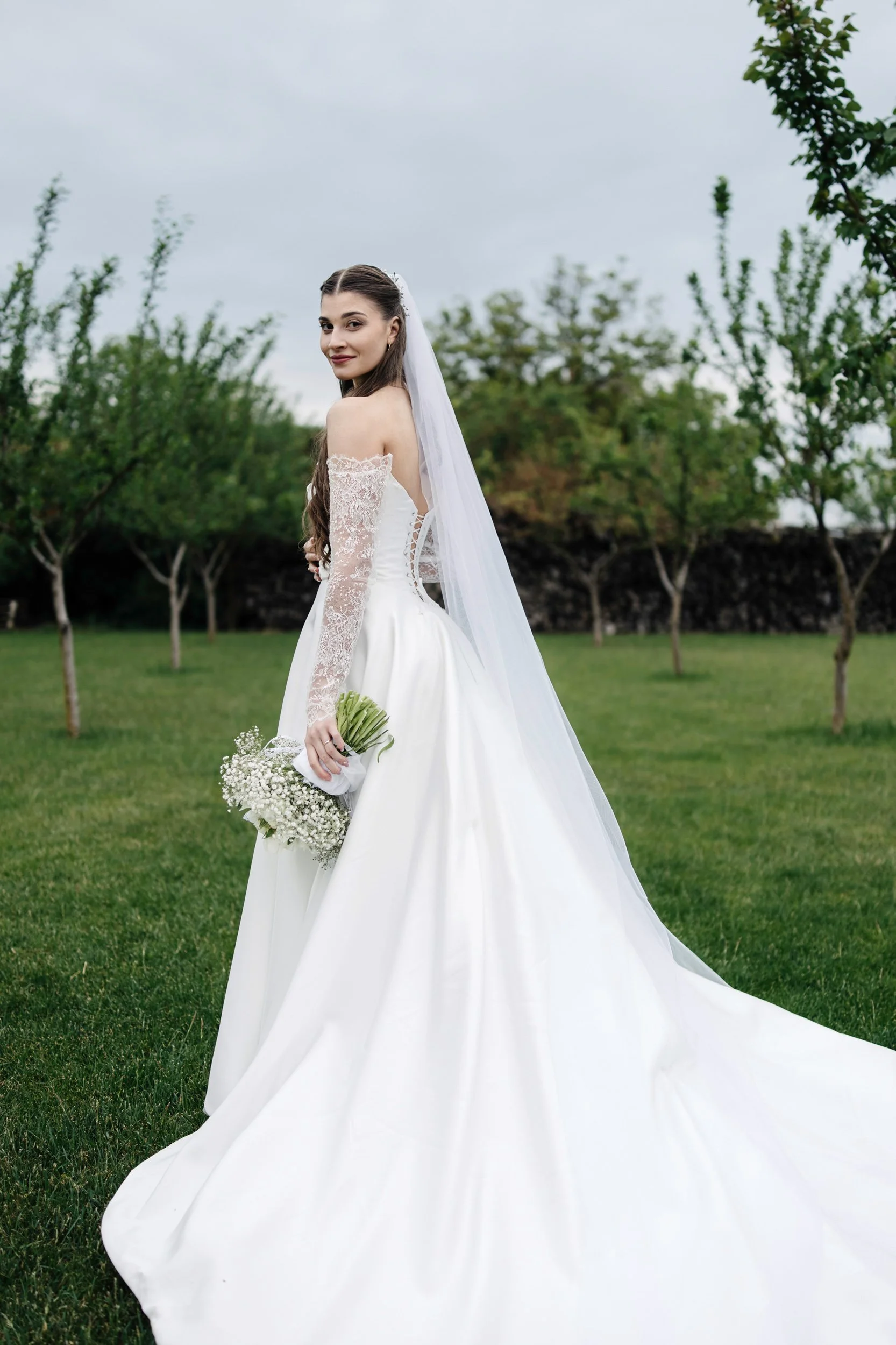 A bride in a white wedding gown with lace sleeves, holding a bouquet of baby's breath flowers, standing on green grass with trees and a stone wall in the background under a cloudy sky.