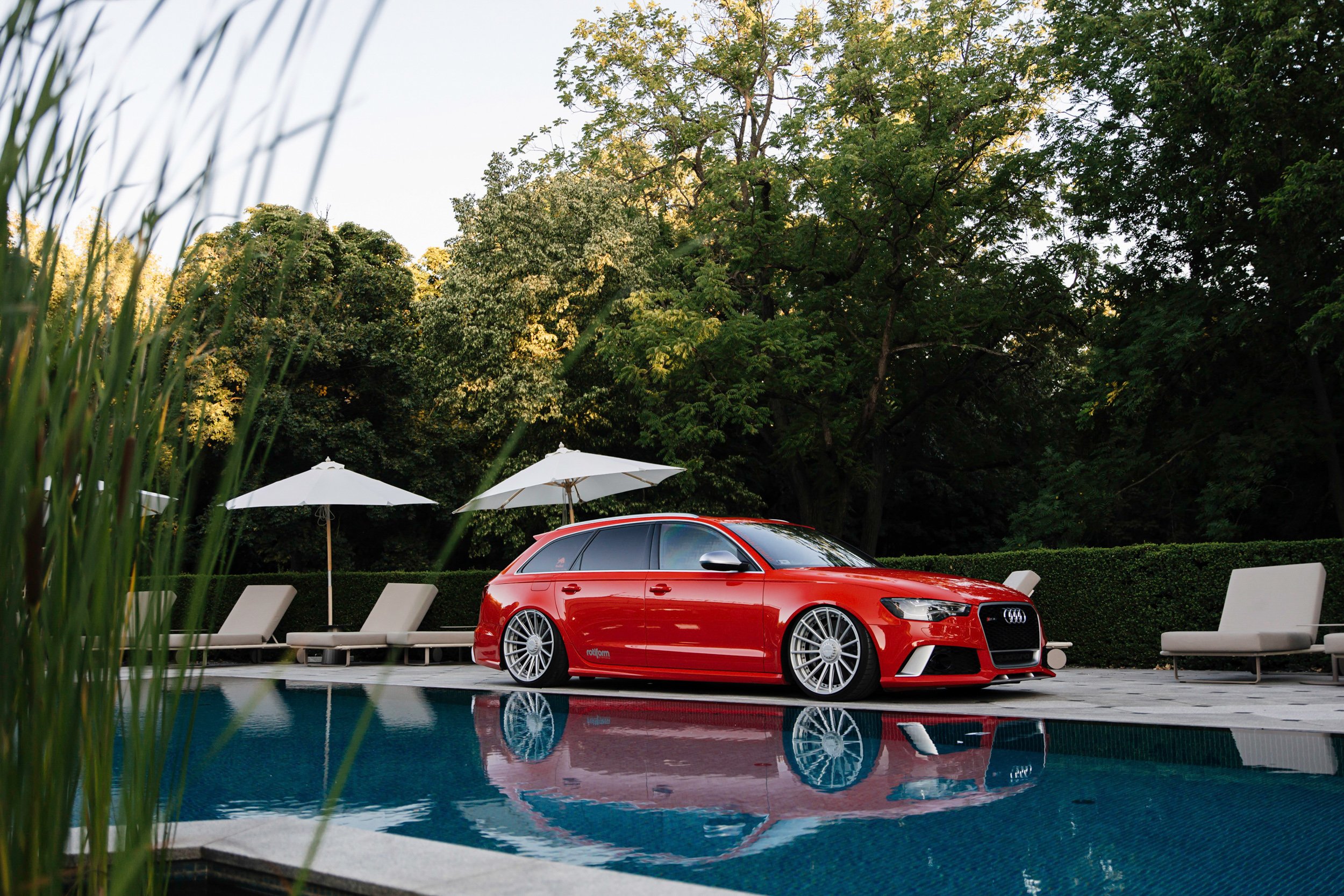 A red Audi car parked by a swimming pool with white lounge chairs and umbrellas, surrounded by green trees and foliage.