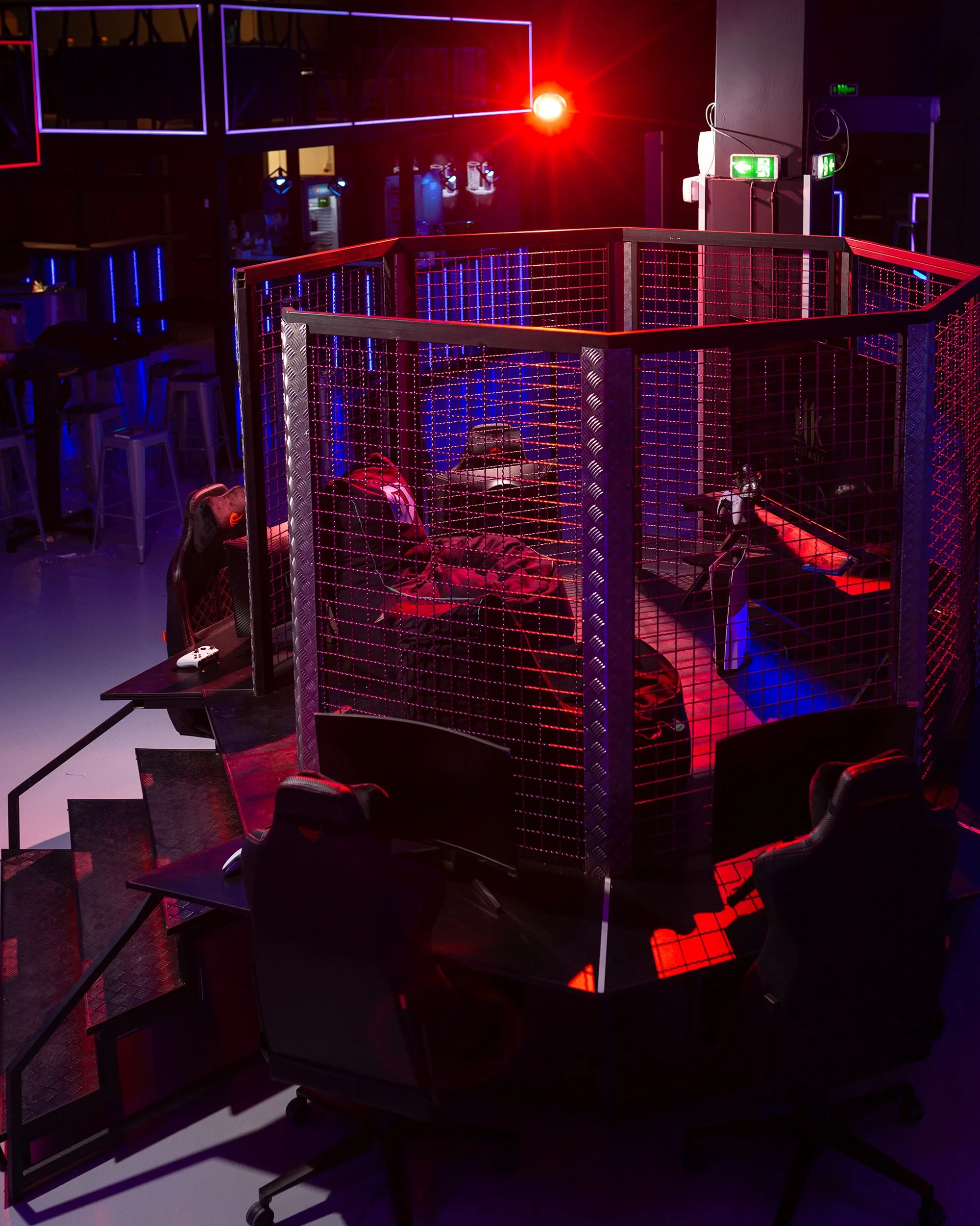 An indoor gaming or entertainment area with neon and LED lights, featuring a fencing enclosure for activities, gaming chairs, and monitors. The space is dimly lit with red, purple, and blue lighting, creating a vibrant atmosphere.