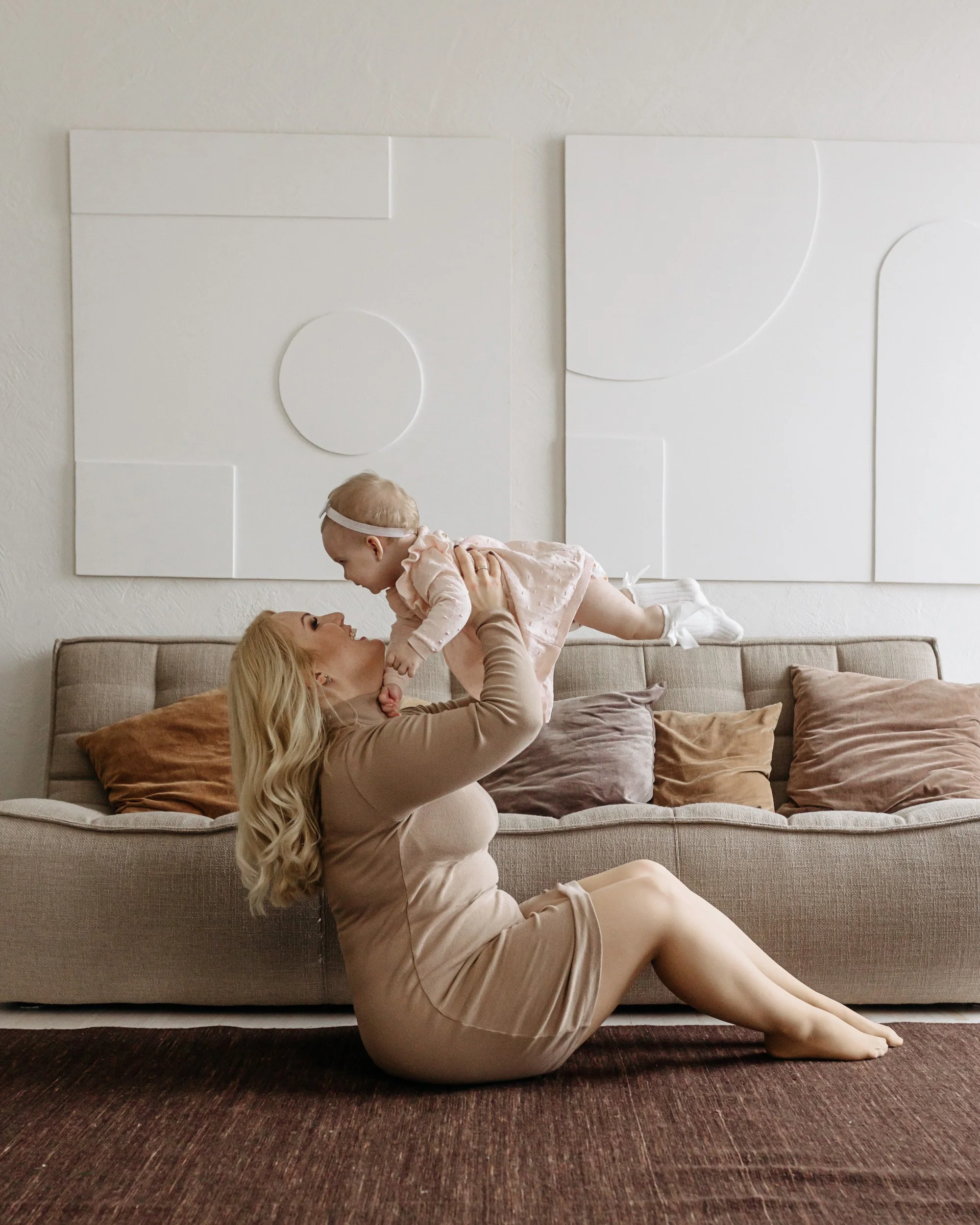 A woman with long blonde hair in a beige dress is sitting on a dark rug, lifting a baby girl with light hair and a headband onto her lap. They are in a room with a beige couch, pillows, and white abstract wall art.