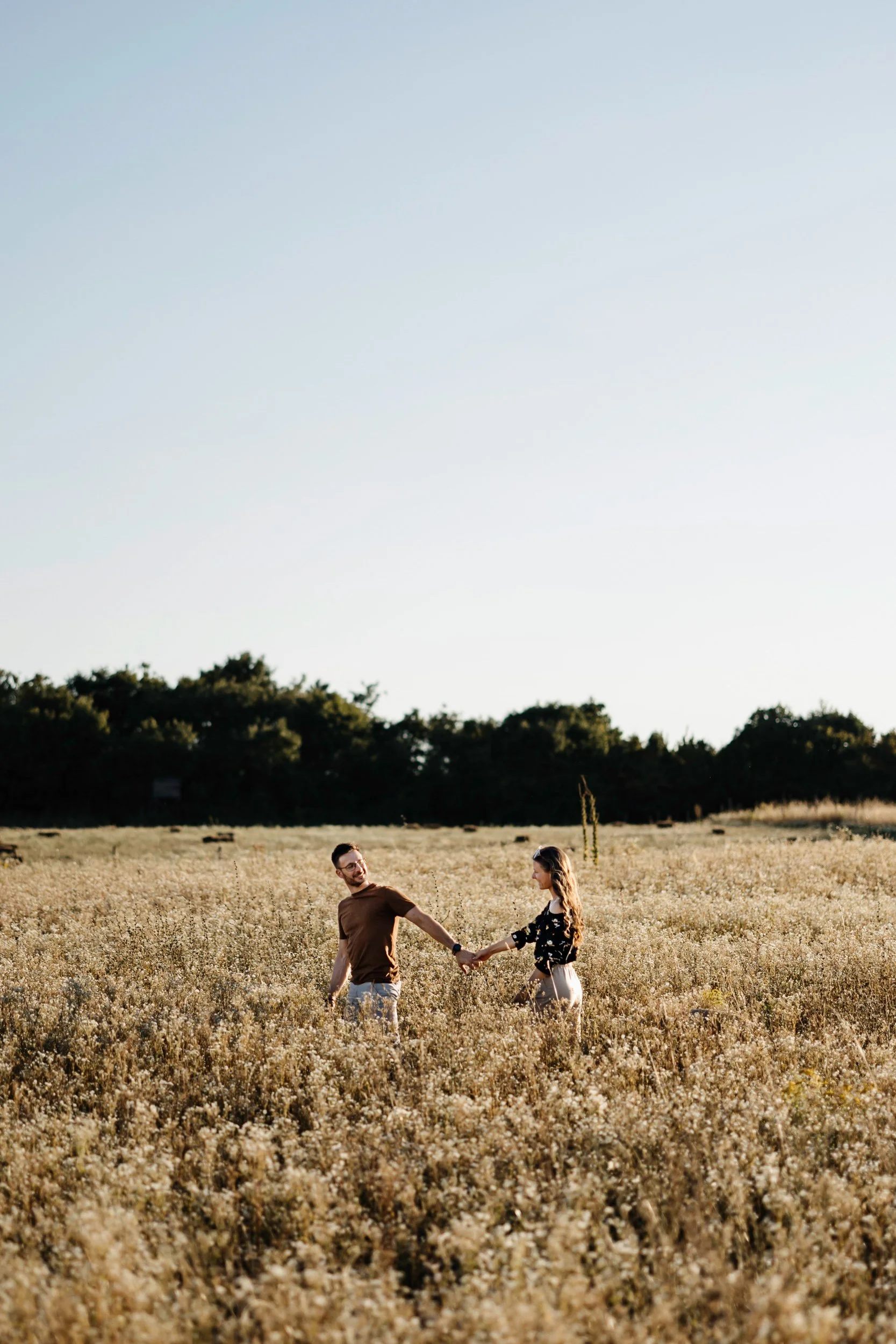 A couple holding hands and smiling in a field of tall grass or wildflowers with a tree line in the distance and a clear sky overhead.