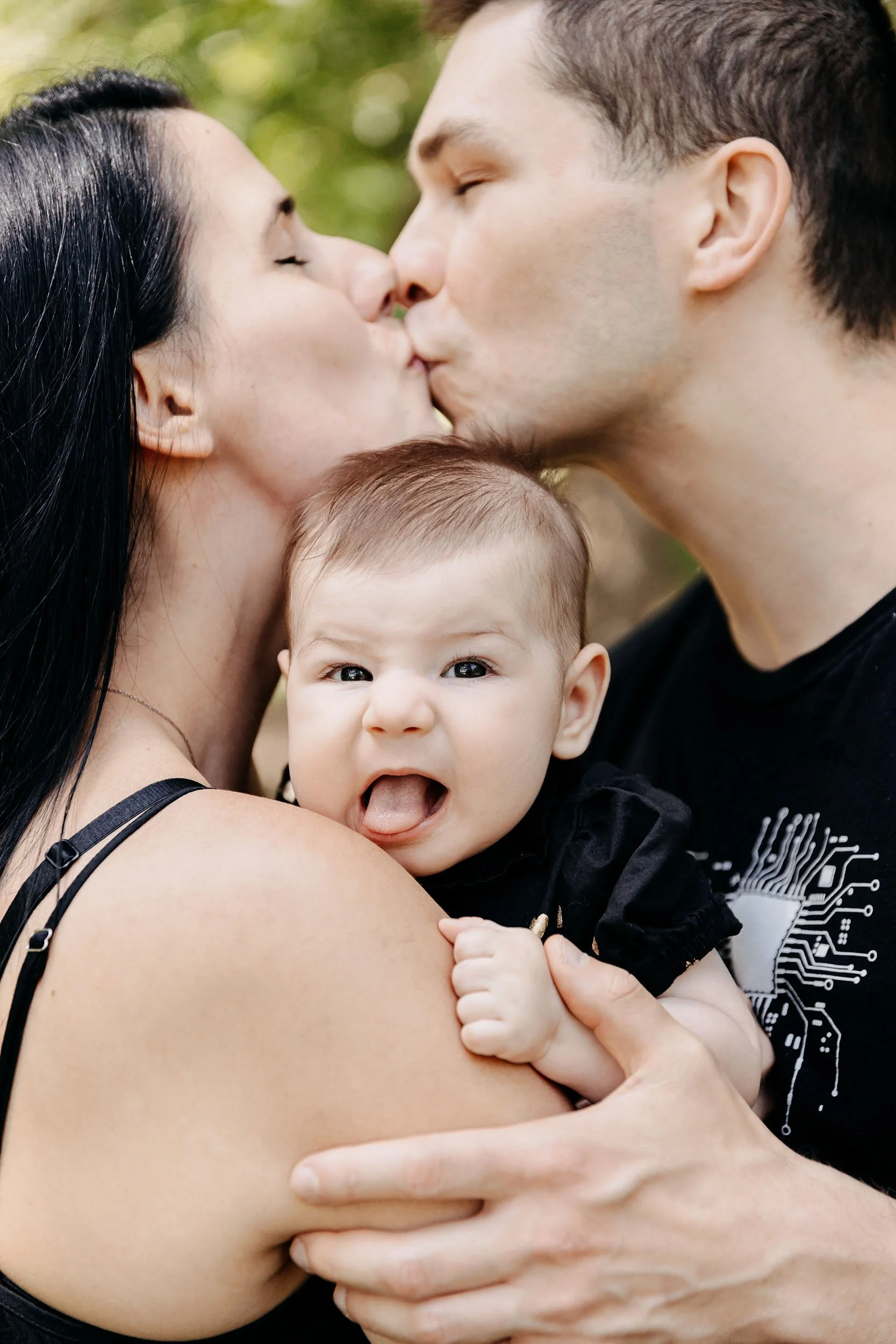 A family of three, a woman, a man, and a baby, sharing a kiss outdoors while the woman holds the baby. The baby is making a playful face with its tongue out.