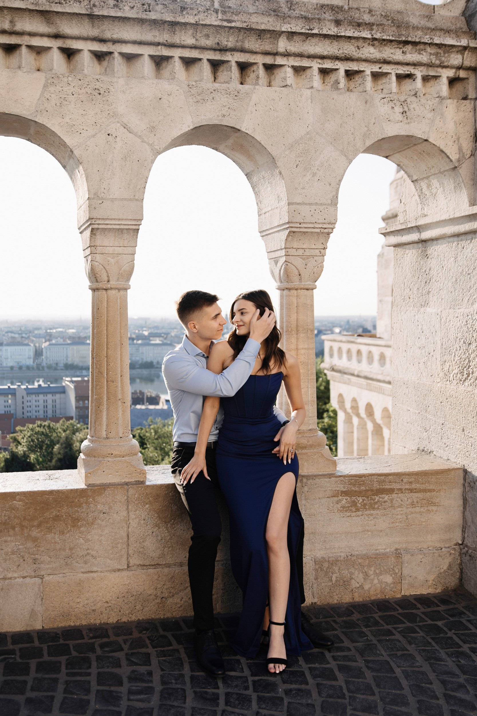 A couple dressed formally, the man wearing a light blue shirt and black pants, the woman in a dark blue strapless gown, standing close together on a stone terrace with arches overlooking a cityscape. The man gently holds the woman's face, and she loo