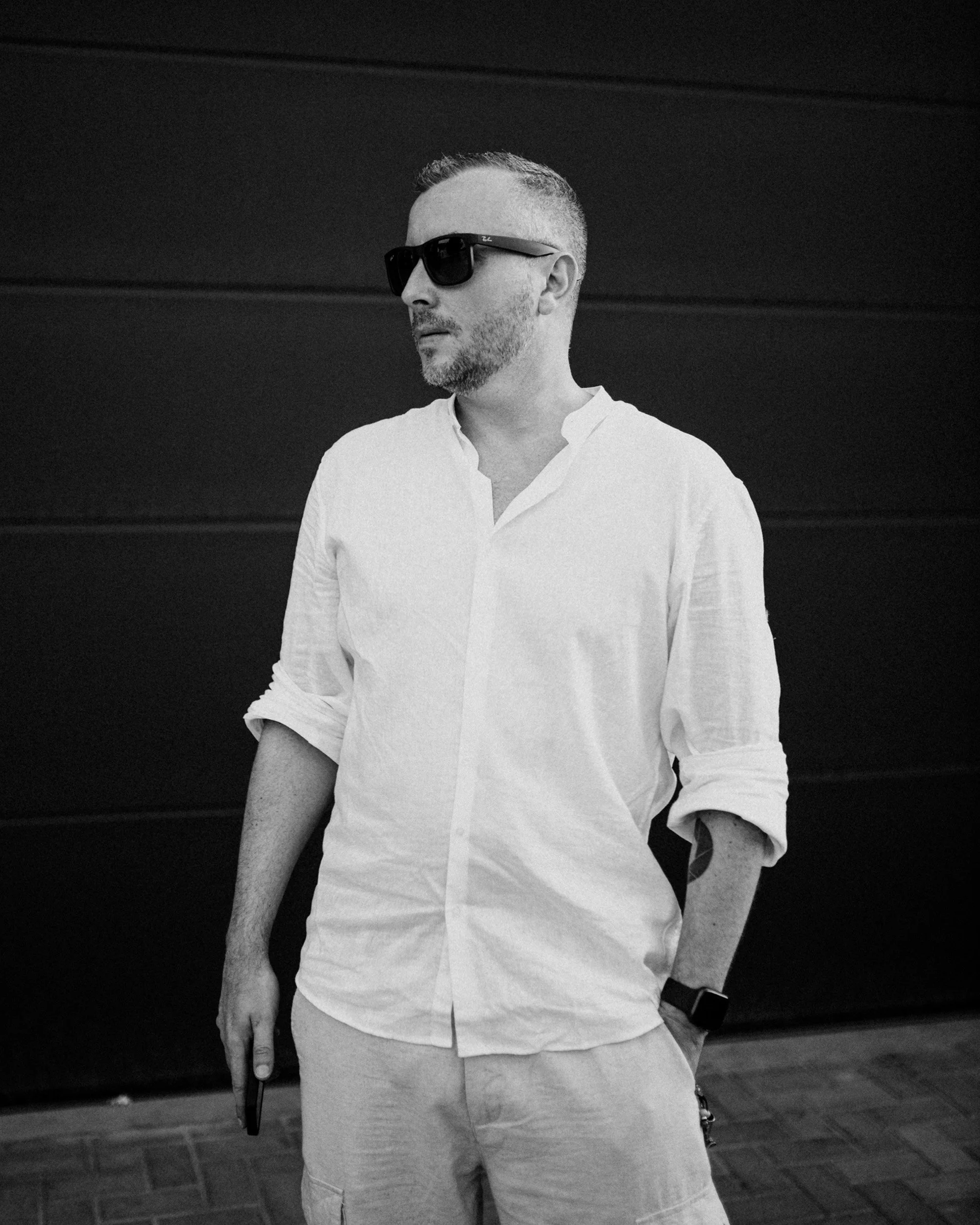 A man wearing sunglasses and a white shirt stands against a dark background. He has short hair and some facial hair, with one hand resting in his pocket and the other holding a cellphone. He is also wearing a watch.