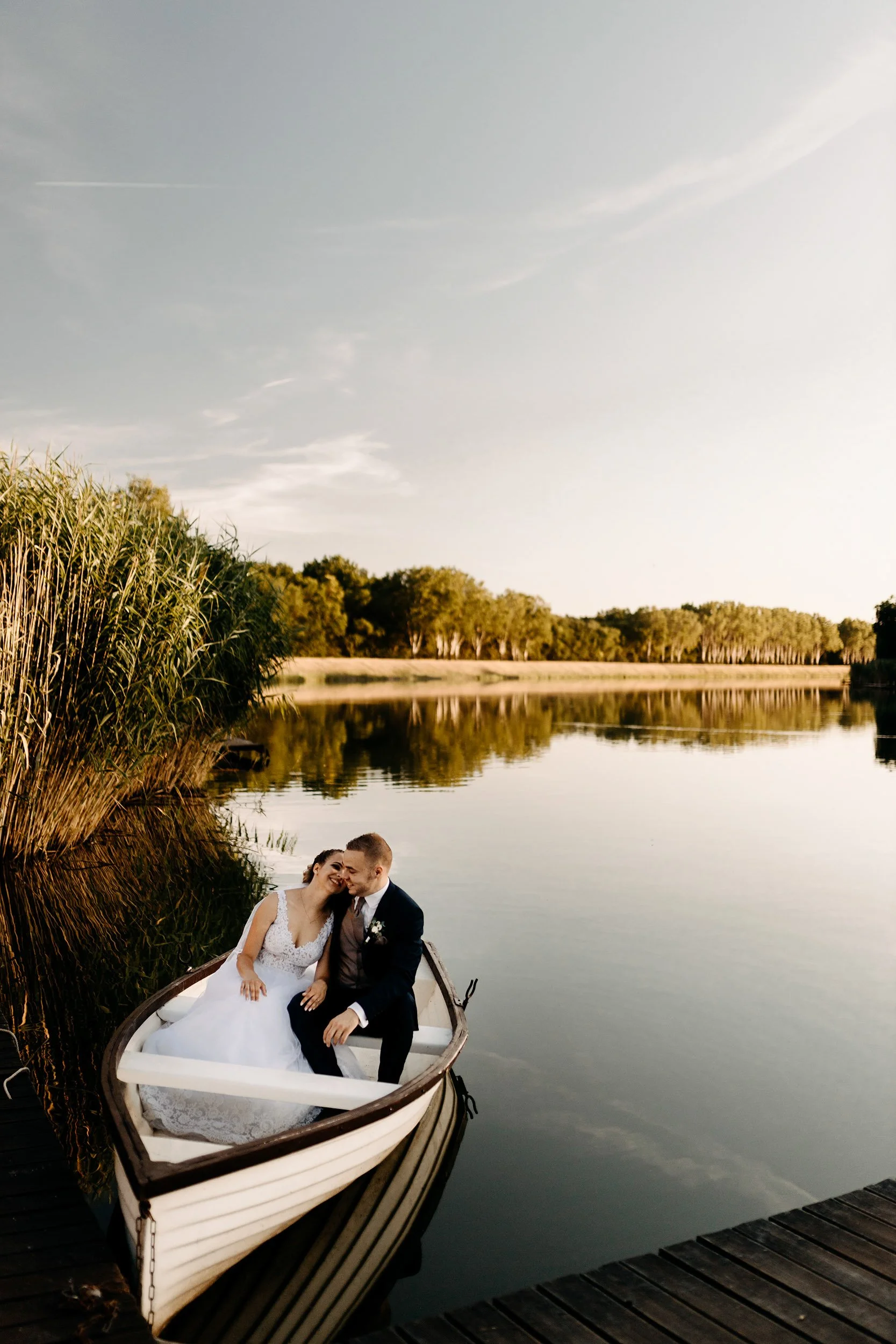 A newlywed couple in wedding attire sitting in a small rowboat on a calm lake with reflections of trees on the water. The bride is smiling and leaning towards the groom, both of them enjoying a joyful moment. The background features lush green trees 