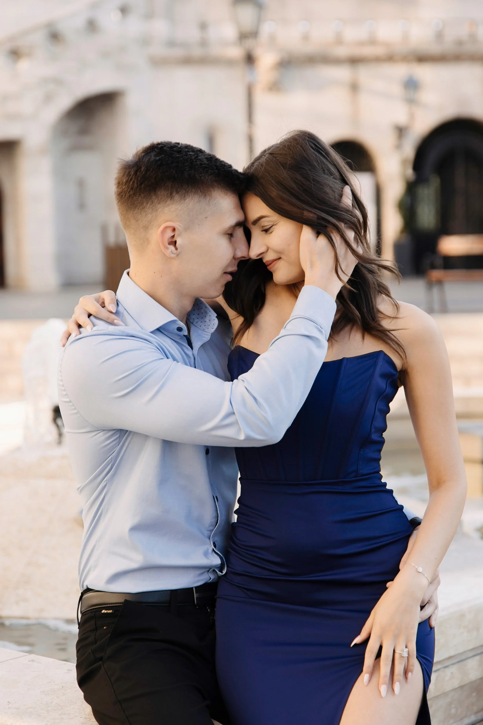 A romantic couple with eyes closed gently touching foreheads and smiling, outside in front of a historic European building with arches and large doorway, during daytime.