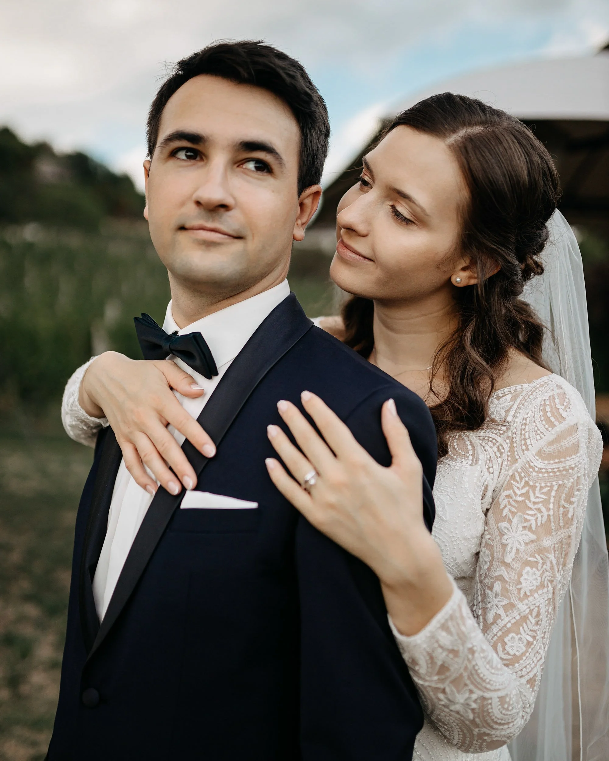 A bride and groom posing outdoors, with the bride standing behind the groom, her arms around his neck, on a cloudy day.
