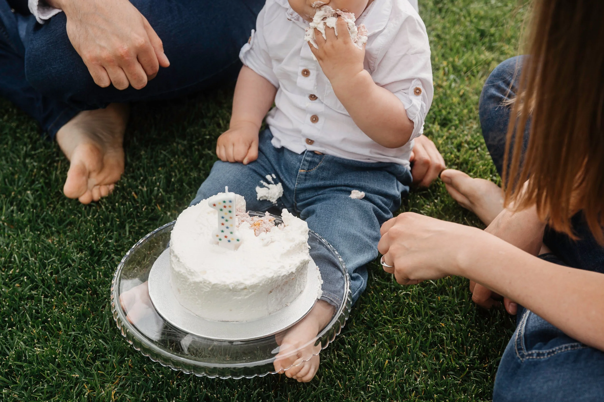 A child sitting on grass eating cake with a birthday candle, surrounded by adults celebrating outdoors.