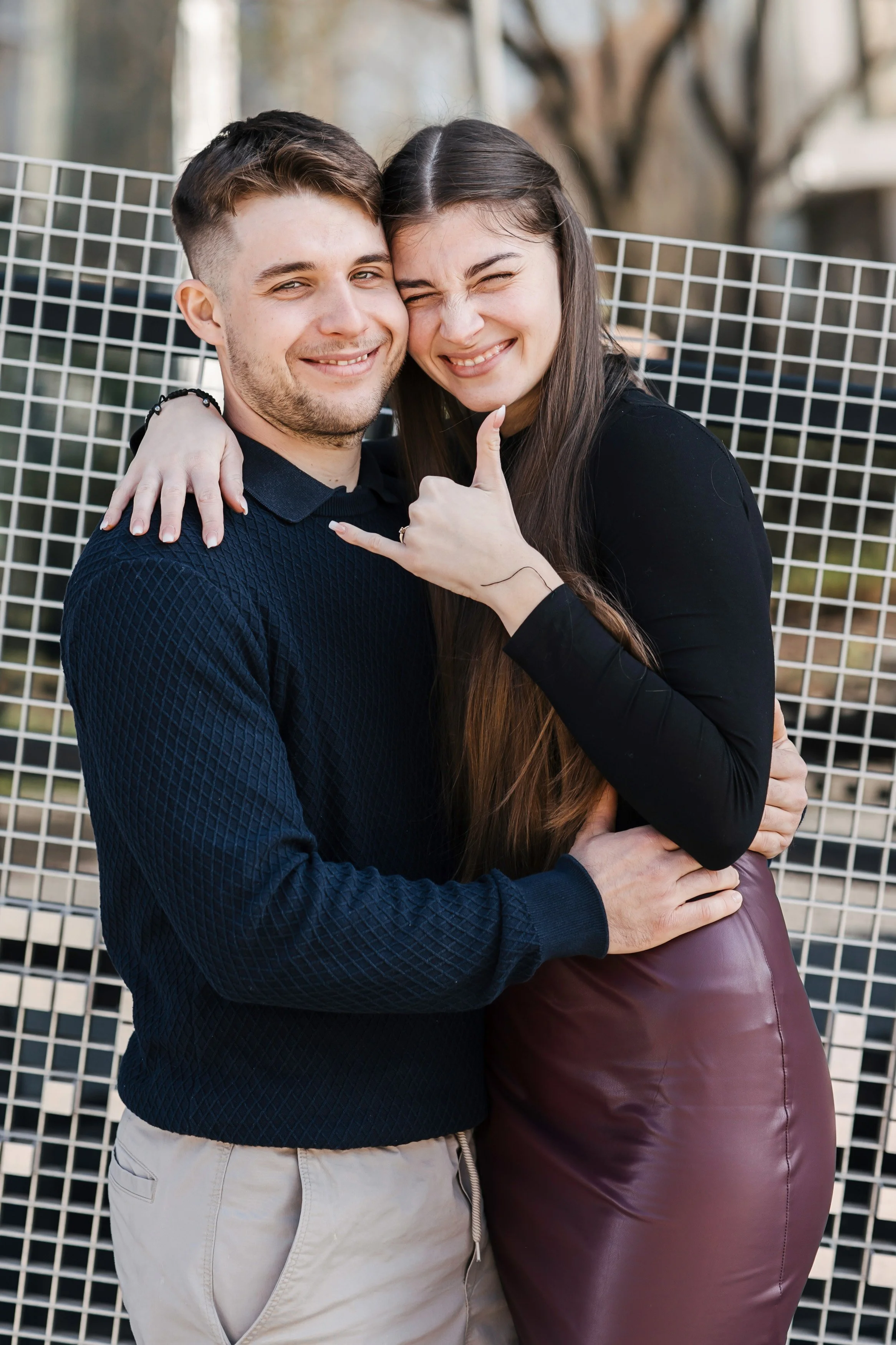A young man and woman are hugging and smiling outdoors, with a white metal fence in the background.
