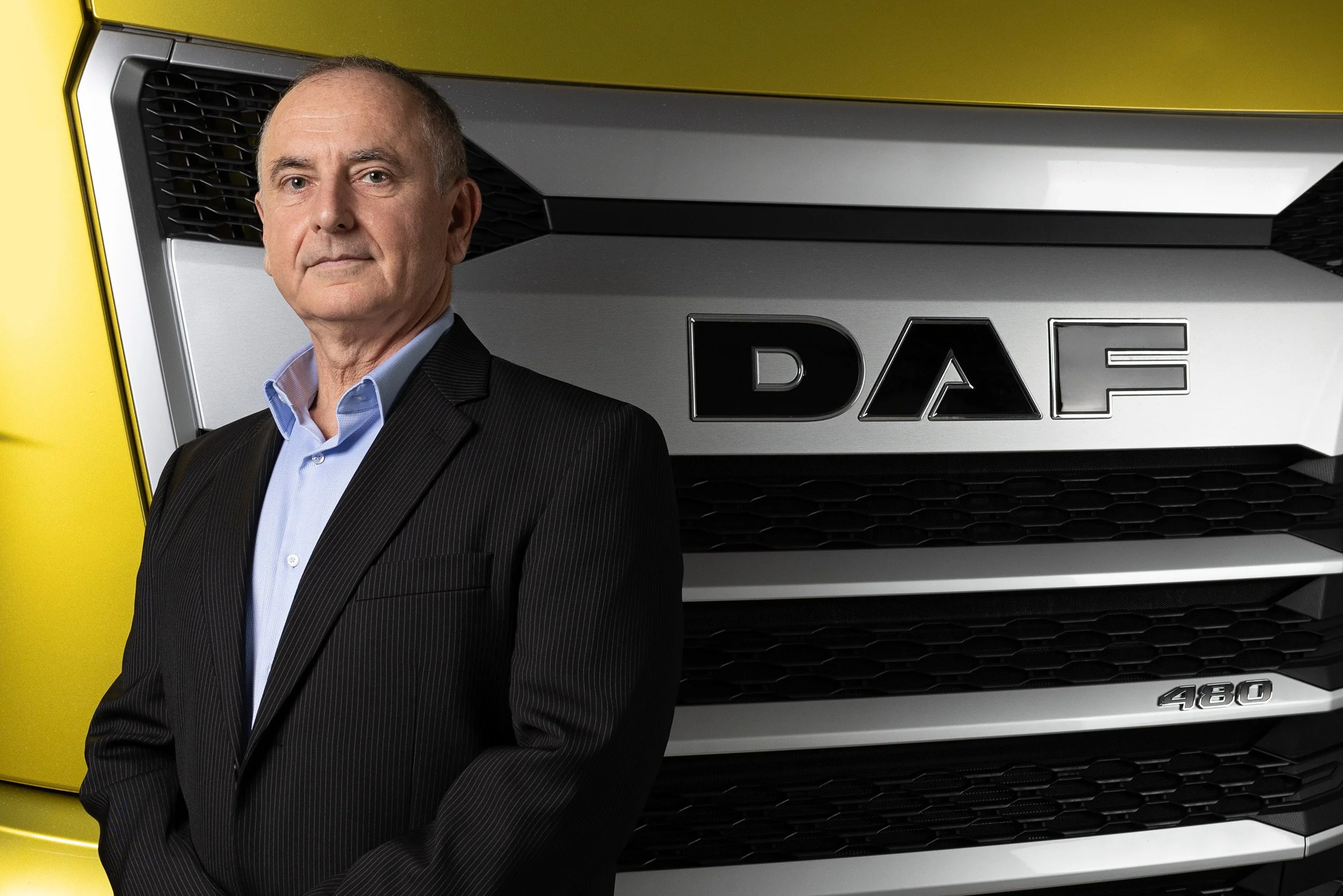 A middle-aged man in a black suit and light blue shirt standing in front of a yellow vehicle truck with a DAF logo on the grille and the number 480 on the lower side of the front bumper.