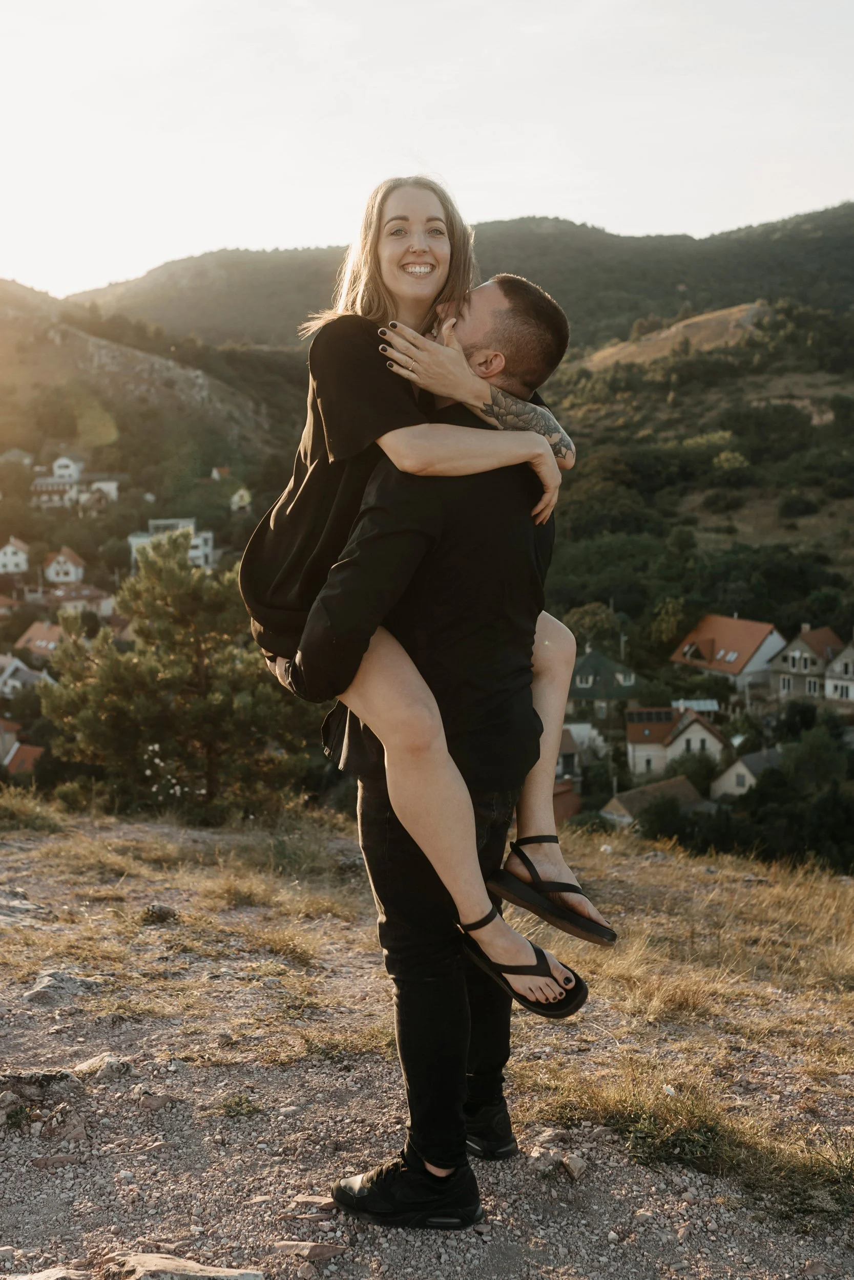 A couple outdoors on a hill, with the woman being carried on the man's back, smiling at the camera during sunset, with houses and mountains in the background.