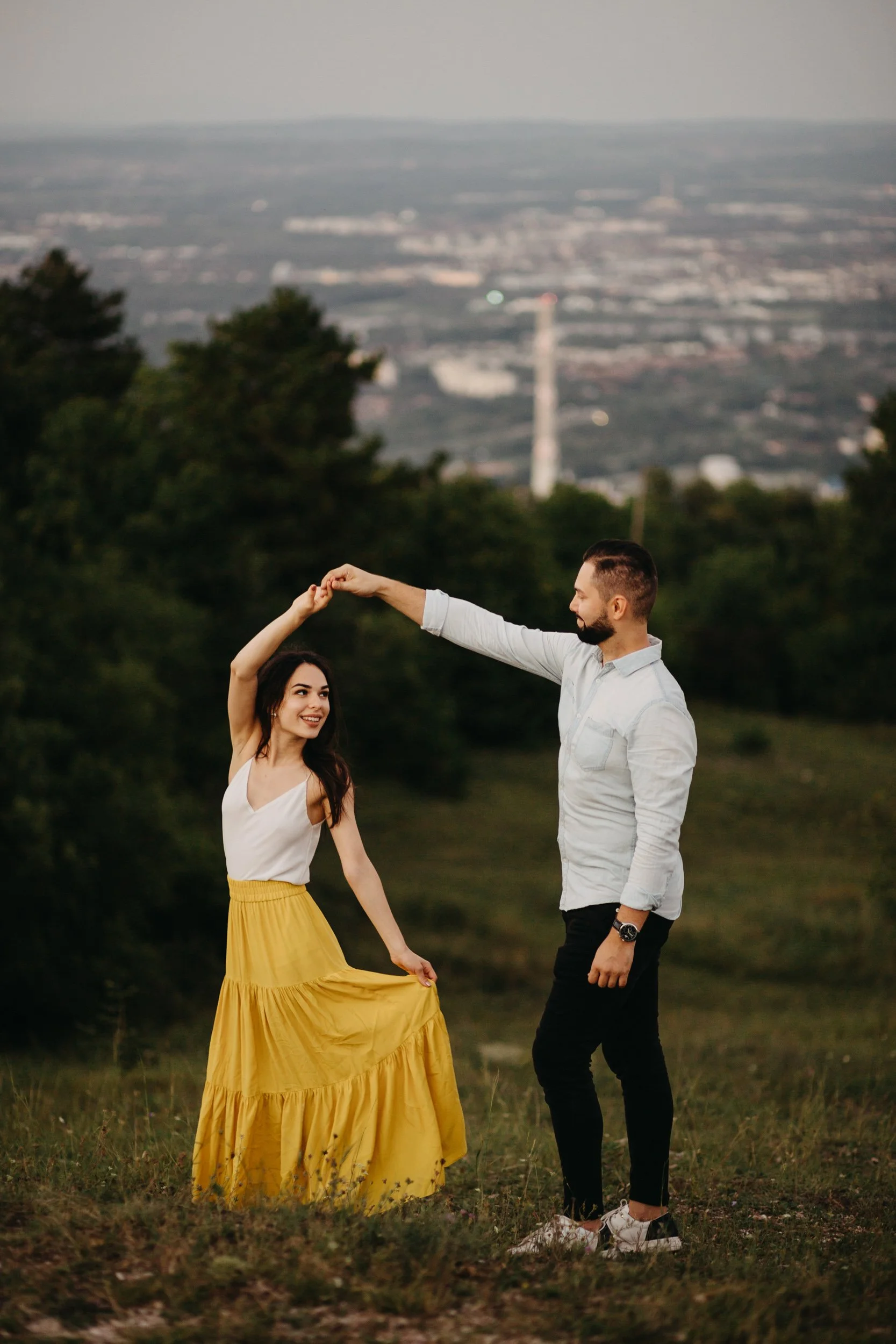 A man and woman are dancing outdoors on a grassy field, with the man holding the woman’s hand as she twirls in a yellow skirt. Trees and a distant city skyline are in the background.