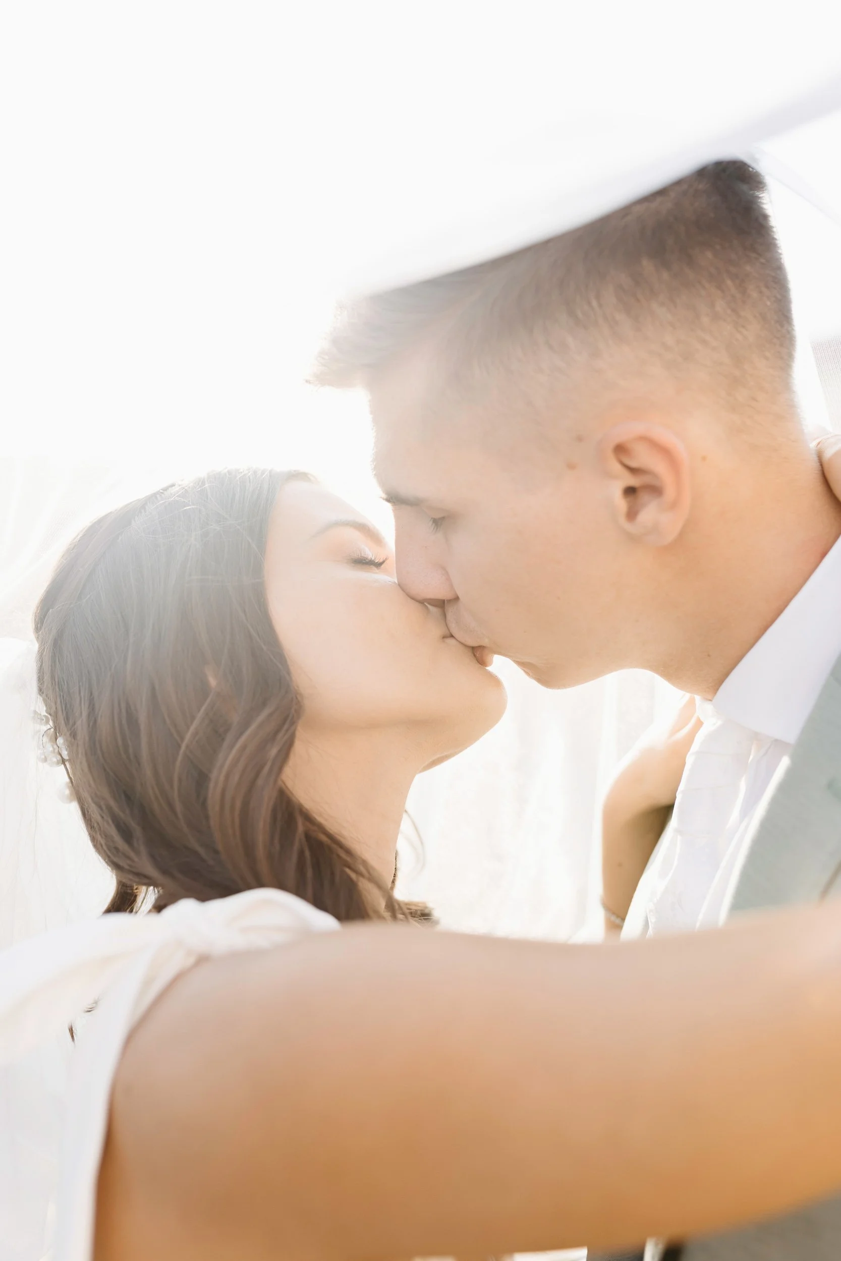 A close-up of a couple sharing a kiss, with the woman having dark, wavy hair and the man with short hair and a fade haircut, both with their eyes closed, in a bright, softly lit setting.