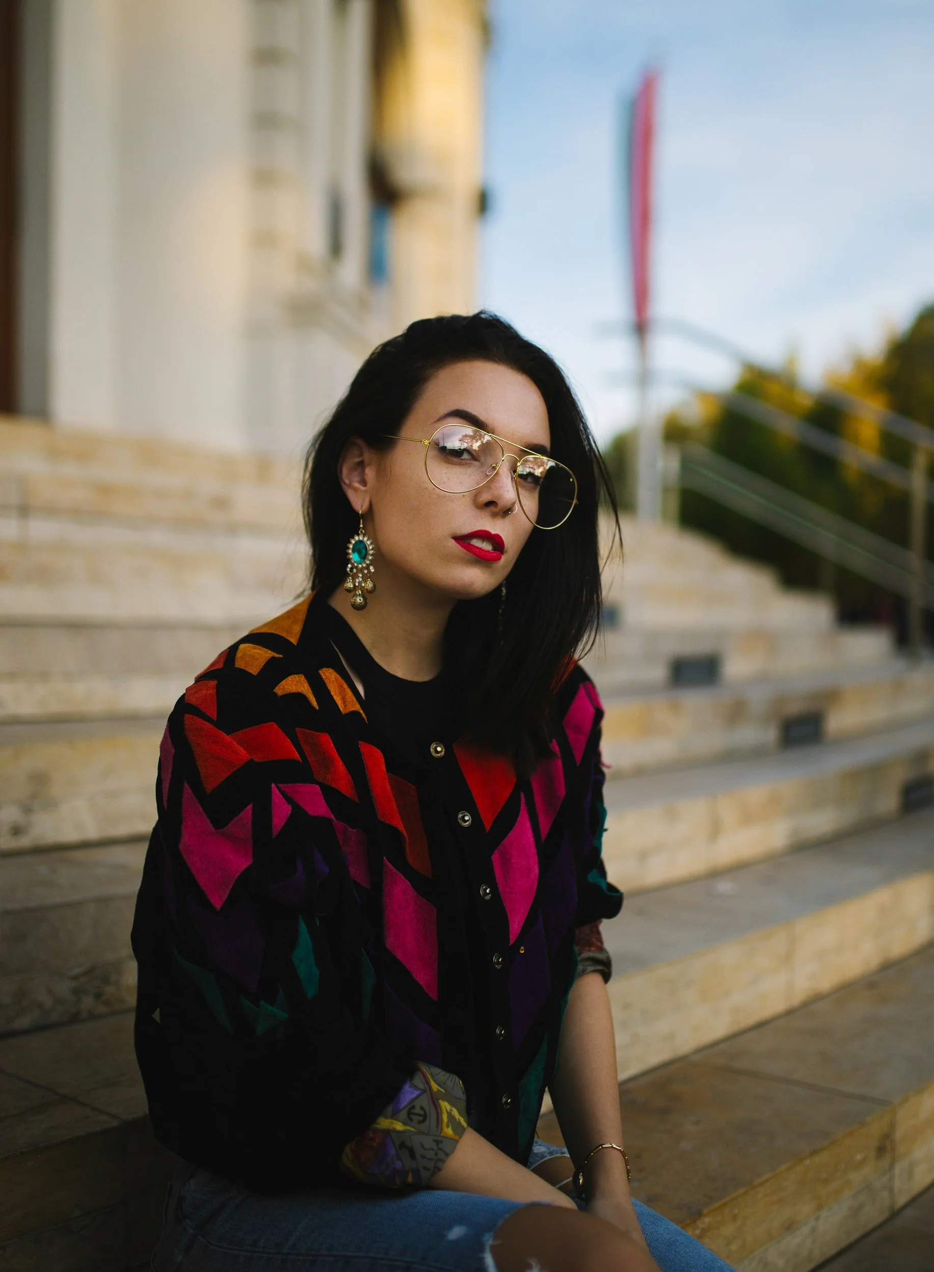 Young woman with dark hair, wearing large sunglasses, red lipstick, and elongated earrings, sitting on outdoor steps with a colorful jacket and ripped jeans, at dusk with a flag in the background.