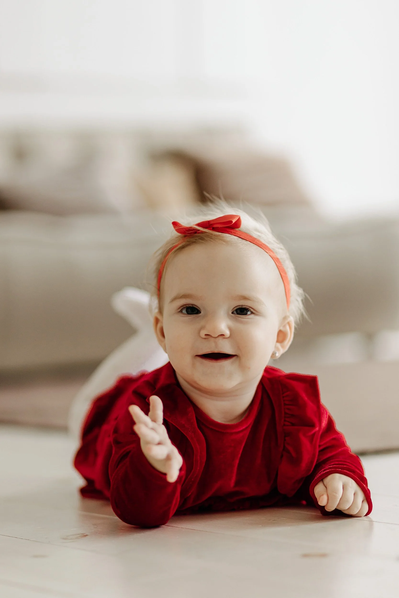 A young girl lying on her stomach on a wooden floor, smiling and waving, wearing a red dress and a red headband.