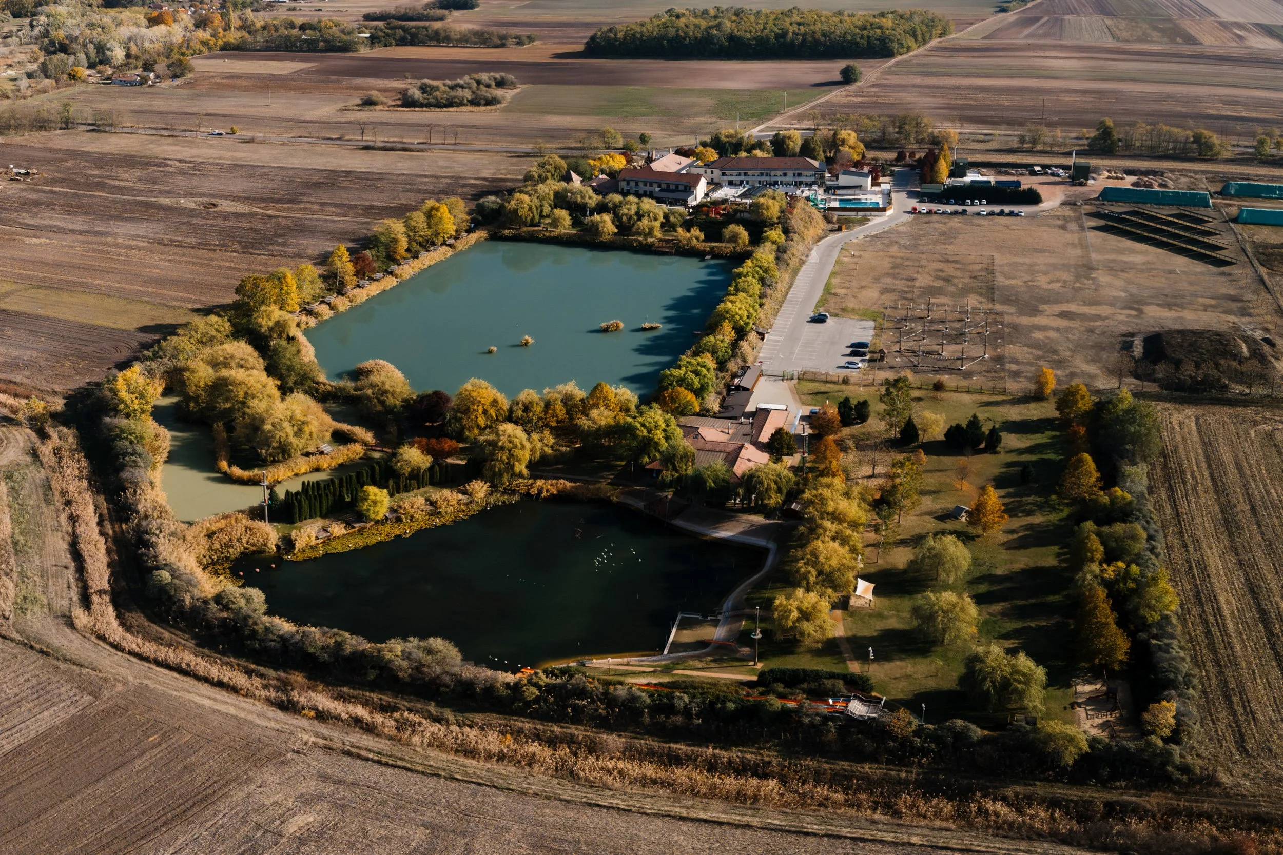 Aerial view of a landscape with a large pond surrounding a complex of buildings, trees, and parking lots, with farmland and fields in the background.
