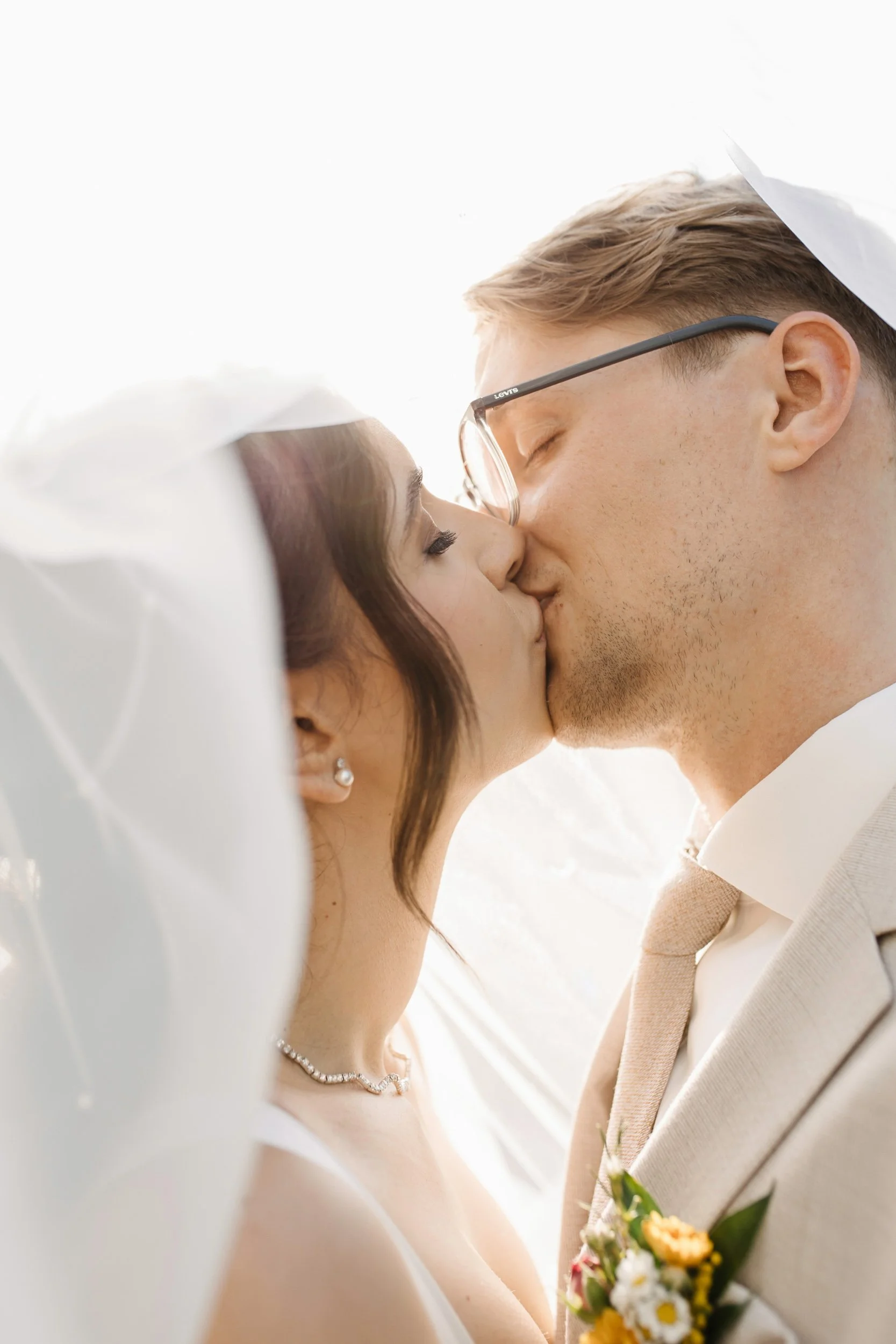 A close-up of a bride and groom kissing during their wedding, with the bride holding a bouquet and wearing a pearl necklace, and both dressed in wedding attire.