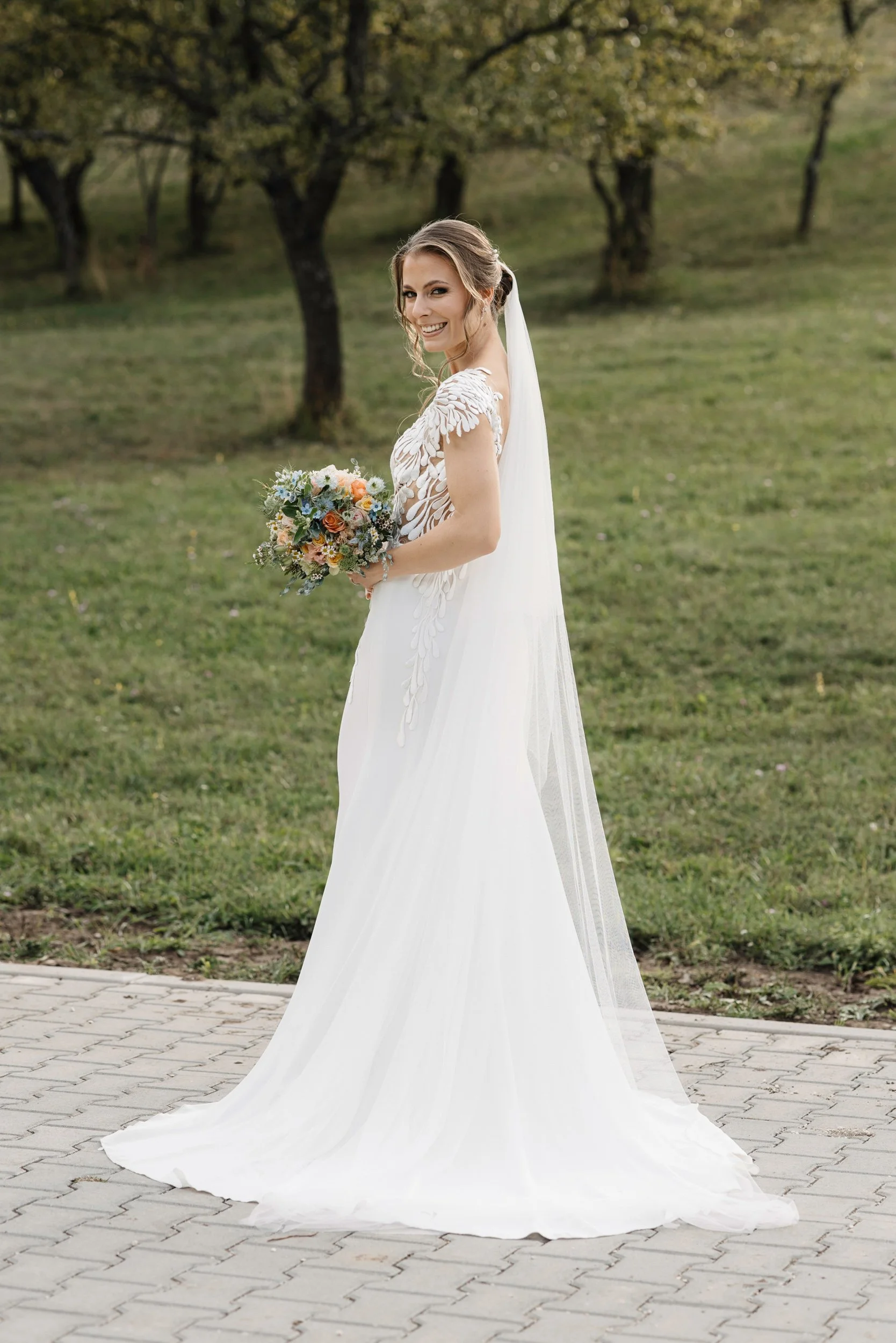 A bride wearing a white wedding dress and veil, holding a bouquet of flowers, standing outdoors on a paved path with trees in the background.