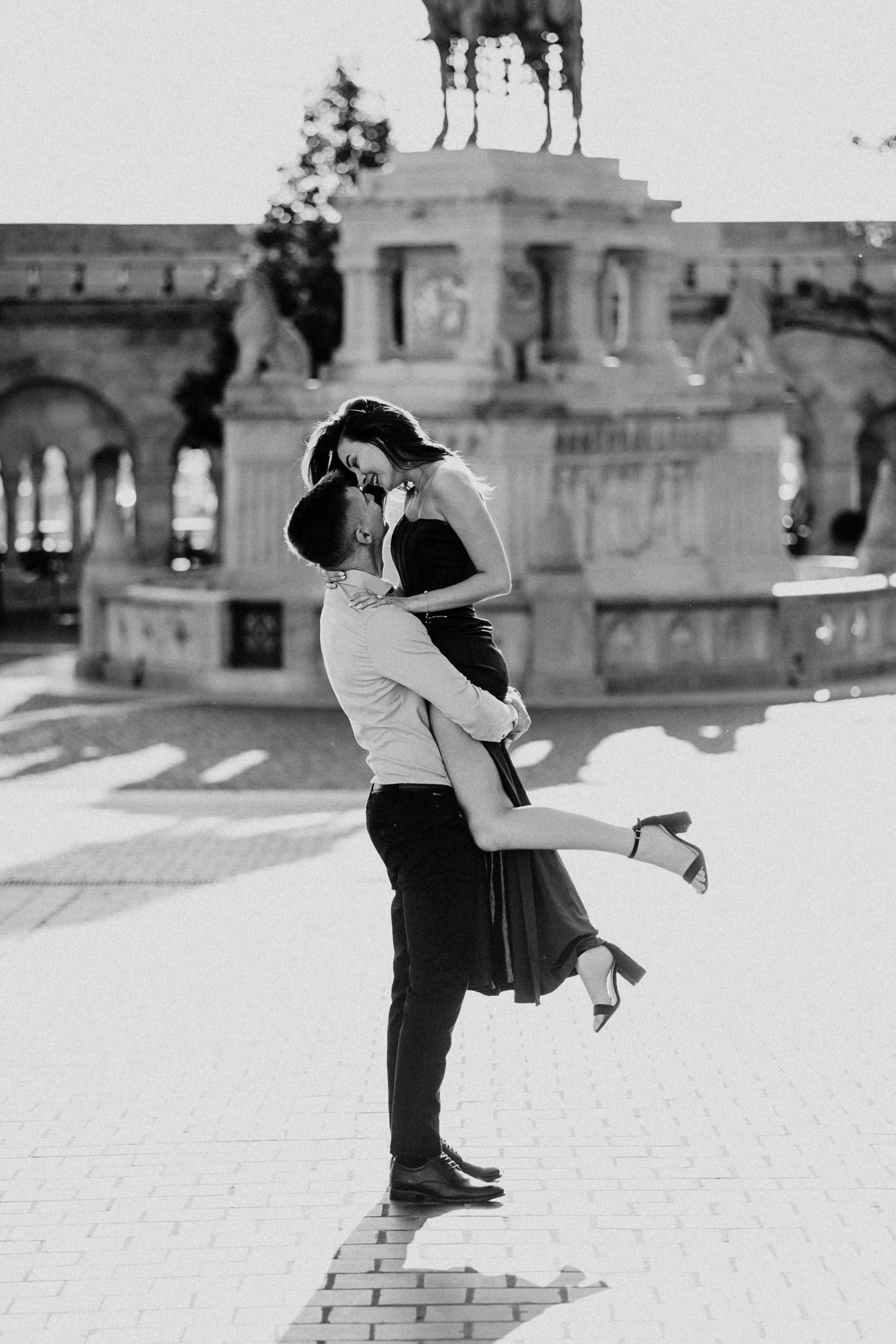 A black and white photo of a man holding a woman who is sitting on his knees, both laughing joyfully in front of a historic monument with statues and ornate architecture.