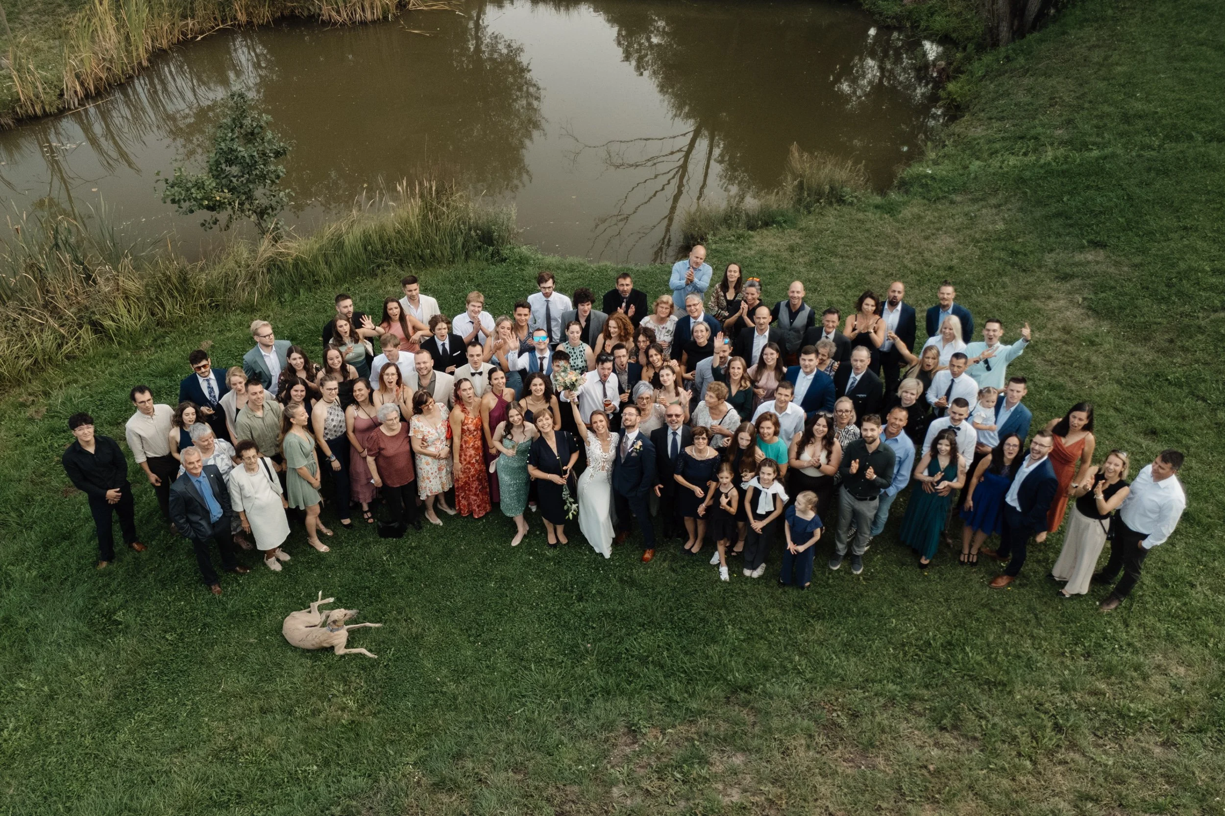 A large group of people gathered outdoors near a pond or small lake, with trees and grass around them. There are men, women, and children dressed in formal and casual attire, smiling and waving at the camera. A dog is lying on the grass in front of t