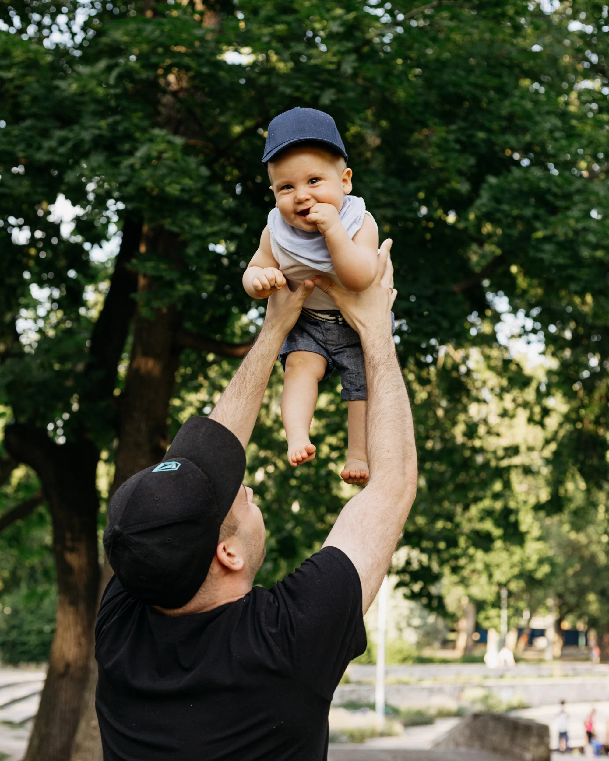 A man wearing a black cap and black shirt is lifting a smiling baby boy wearing a navy cap and face towel outdoors in a park with green trees in the background.
