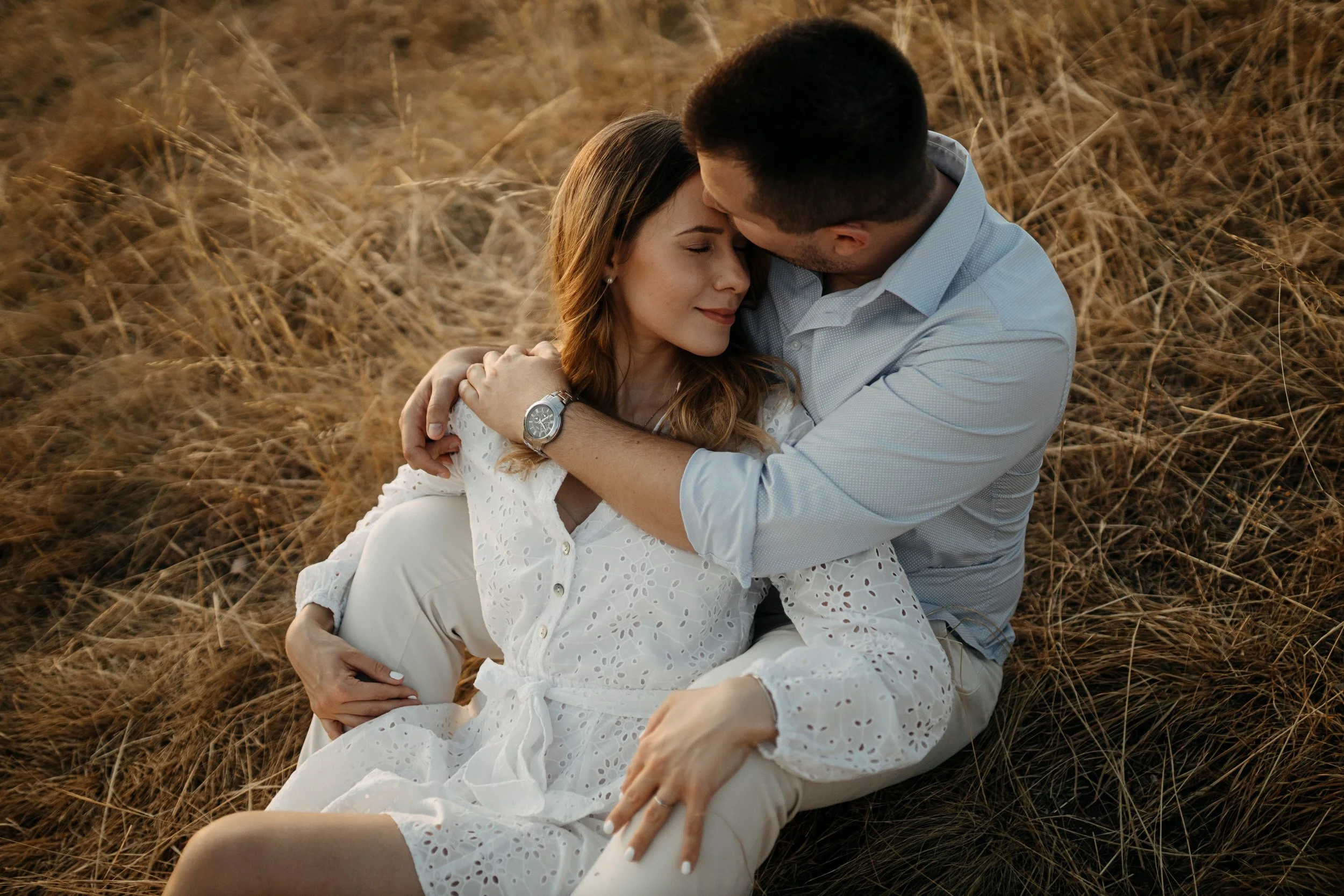 A couple sitting in a field of dry grass, embracing each other closely with their eyes closed, sharing an intimate moment.